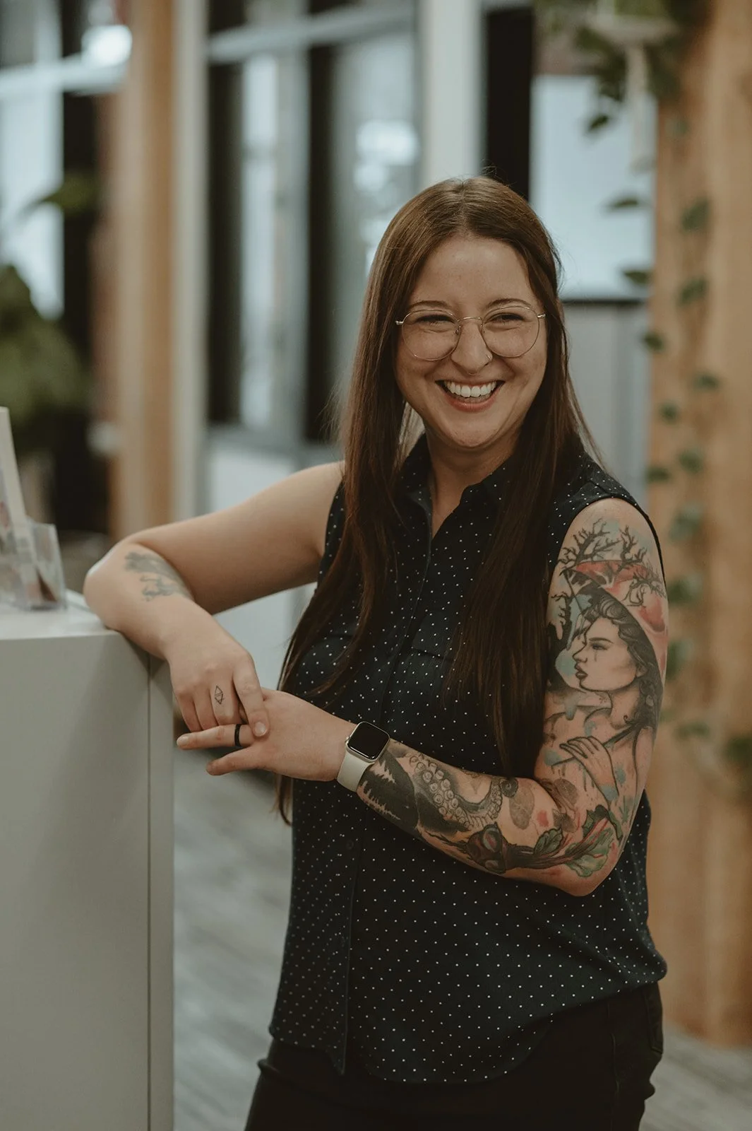 A smiling woman with glasses and long brown hair, wearing a black sleeveless polka dot top, standing indoors with her arms crossed on a white counter. She has tattoos on her left arm and is wearing a smartwatch.