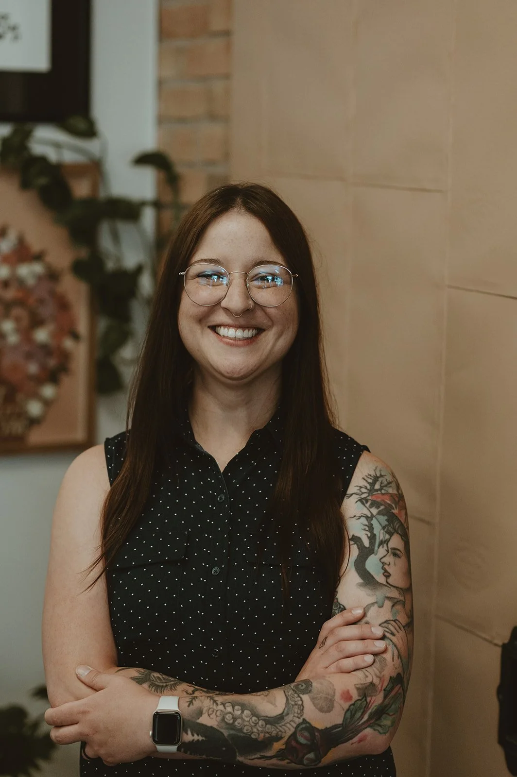 A young woman with long brown hair and glasses smiling, standing with her arms crossed, showing tattoos on her right arm, in an indoor setting with a brick and wooden wall background.