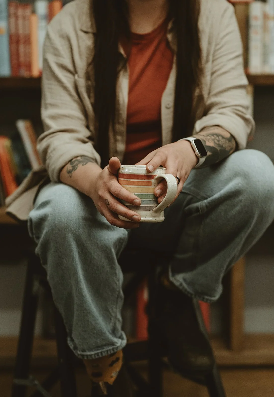 Person sitting on a chair in a library holding a colorful striped mug, wearing a beige jacket, red shirt, and gray jeans.