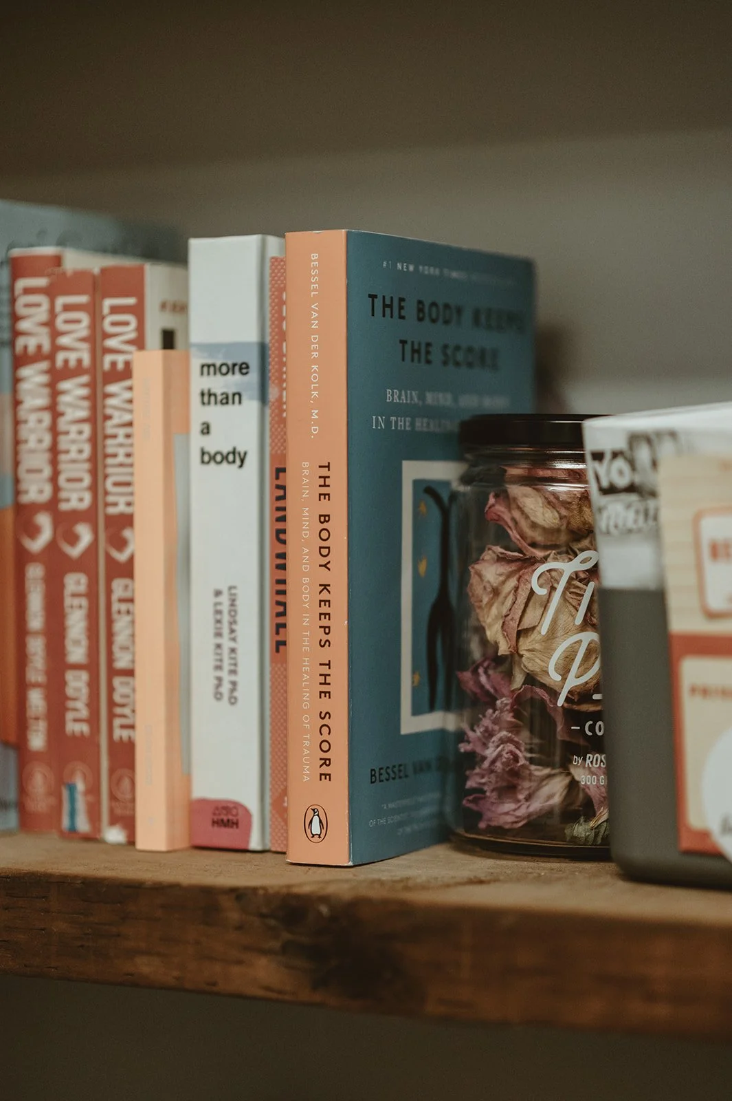 Books on a wooden shelf, including titles about the body and health, such as "The Body Keeps the Score" and "More Than a Body," along with a jar filled with dried flowers or herbs.