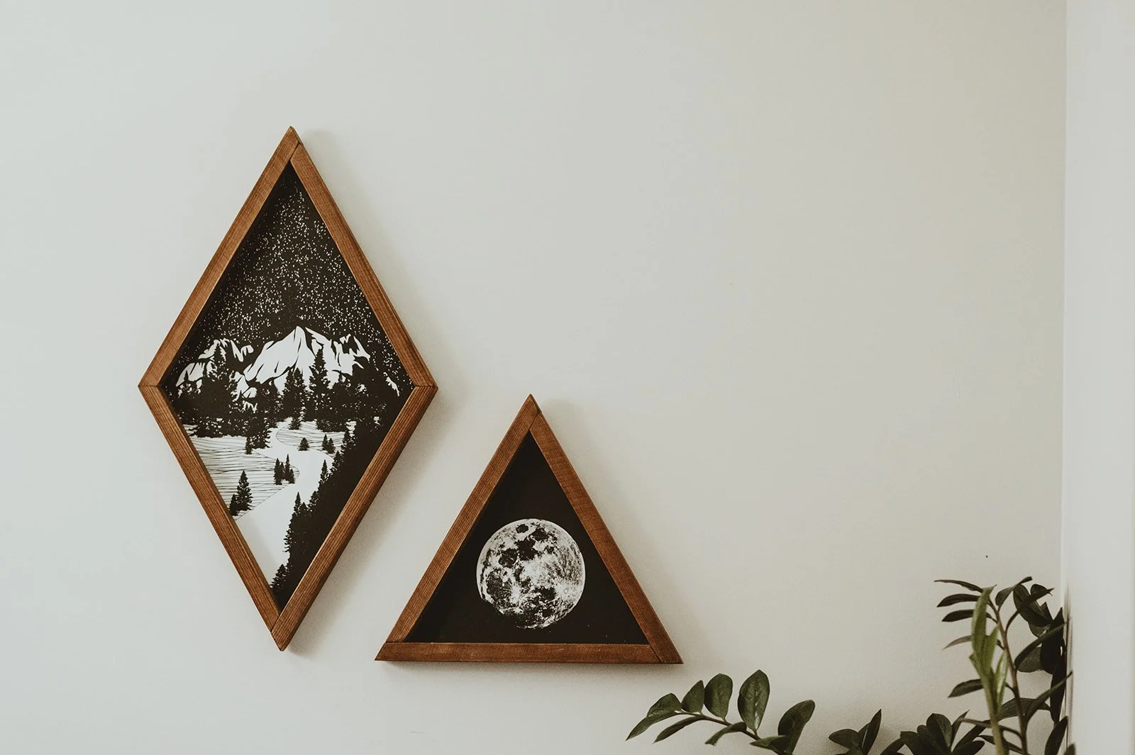 Two diamond-shaped framed art pieces on a white wall, one depicting a mountain landscape with trees and a starry sky, the other showing a full moon, with greenery in the lower right corner.