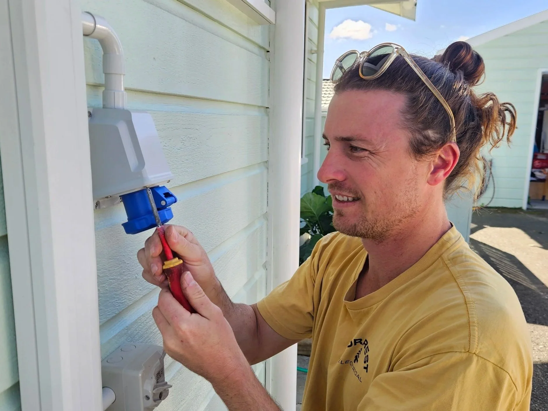 A man wearing glasses on his head is installing or repairing an outdoor electrical outlet or sky camera on the side of a house with green siding. He is using a screwdriver and smiling.