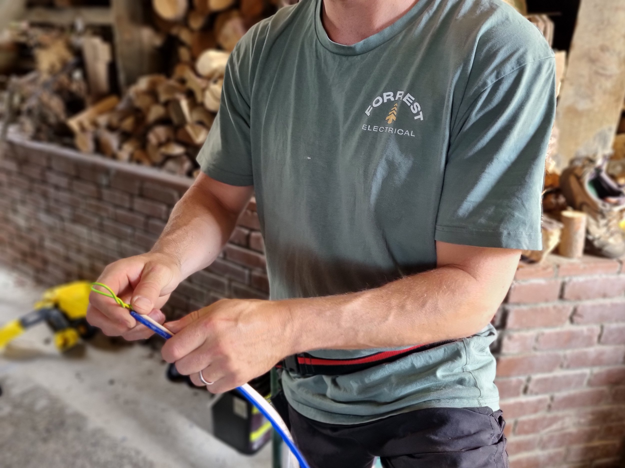 Person wearing a green t-shirt with a logo that says 'Foreest Electrical' and an arrow symbol, working on an electrical project with tools in a setting with stacked firewood and brick wall.