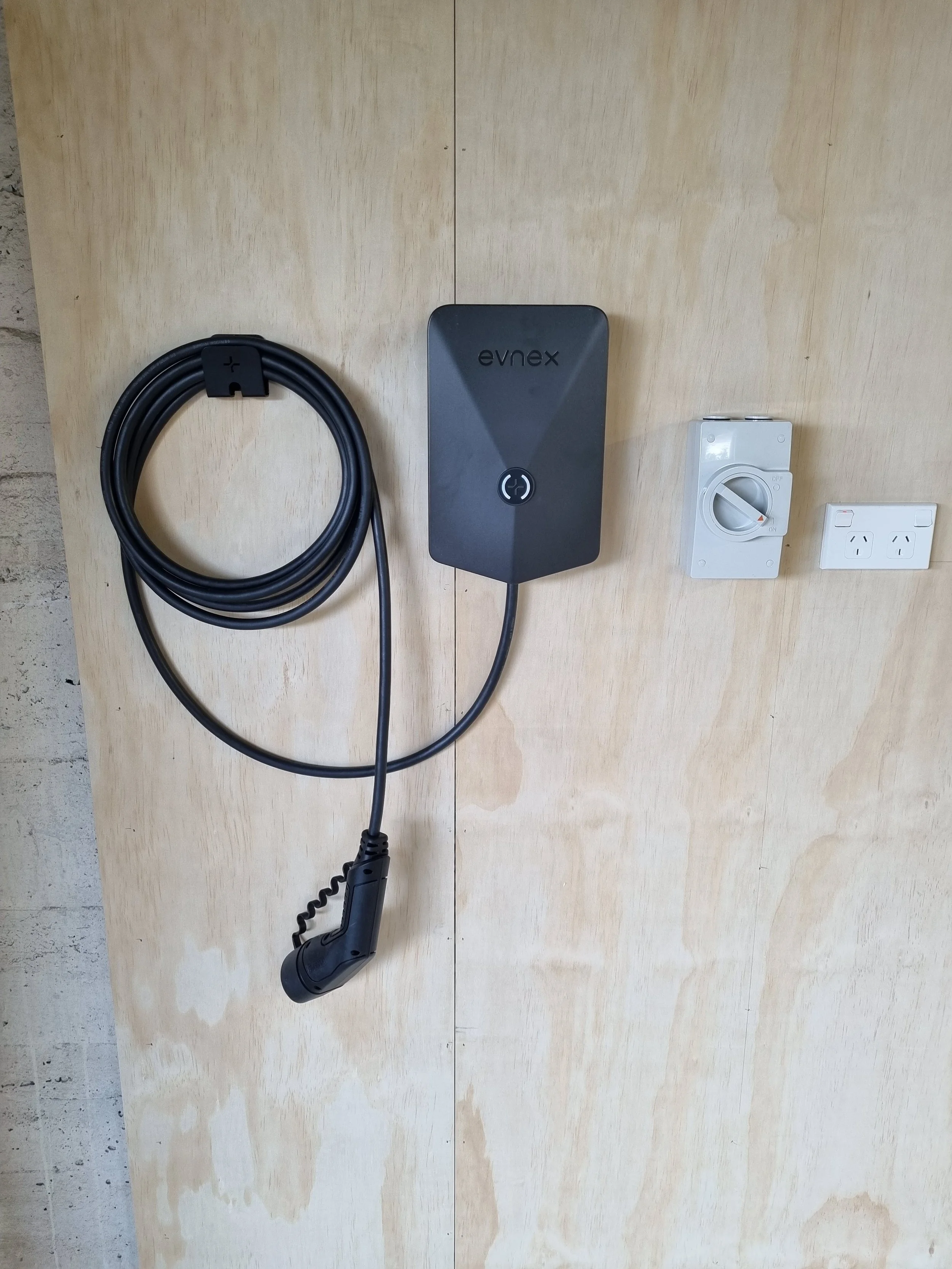 A black electric vehicle charging cable with a plug, a wall-mounted EV charging station labeled 'evnex', a white power outlet, and a white switch mounted on a light wood surface.
