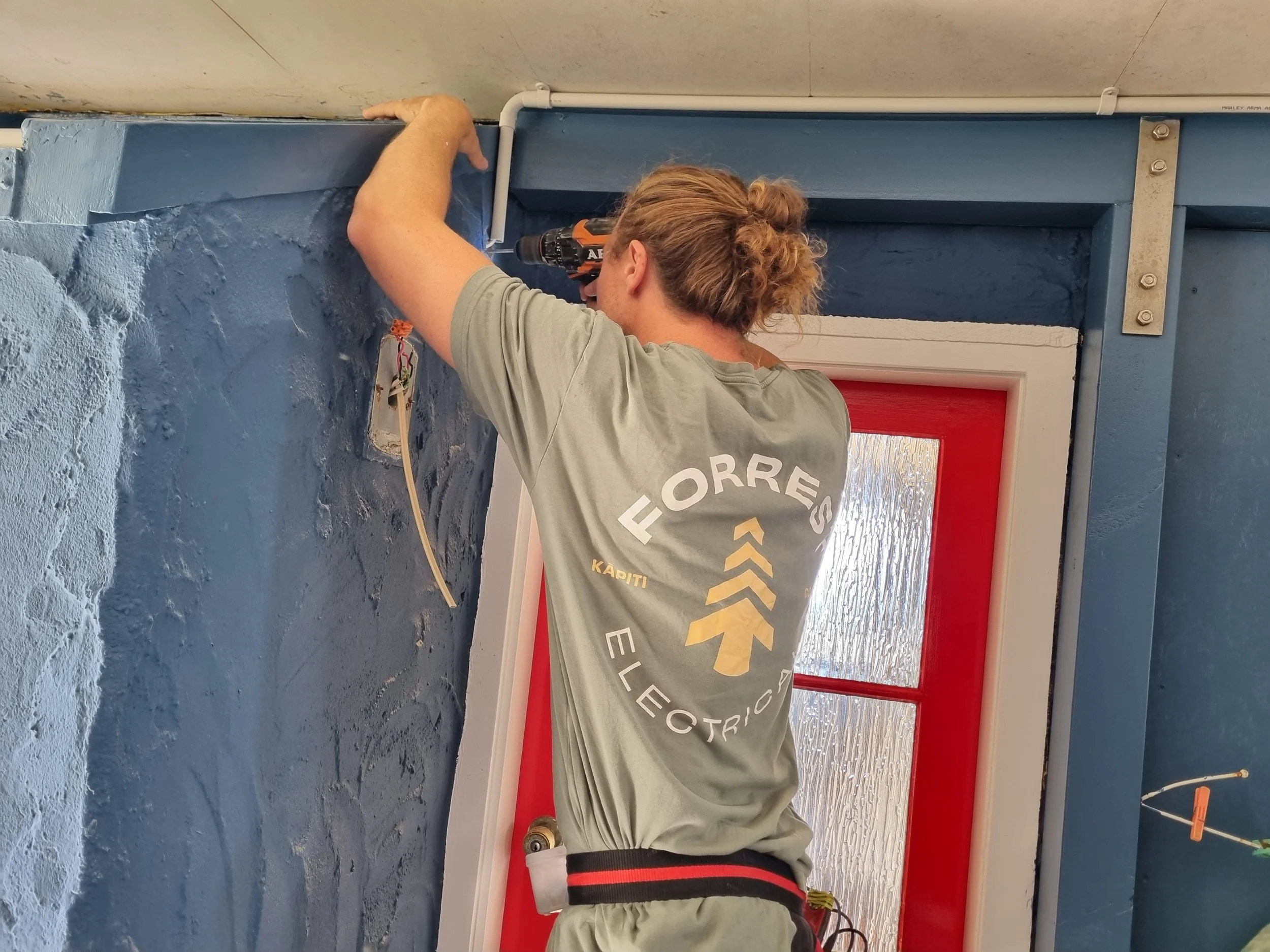 A person with curly hair fixing or installing electrical wiring on a blue wall near a white pipe, wearing a grey shirt with text and a black and red waistband, in a room with a red door and textured grey wall.