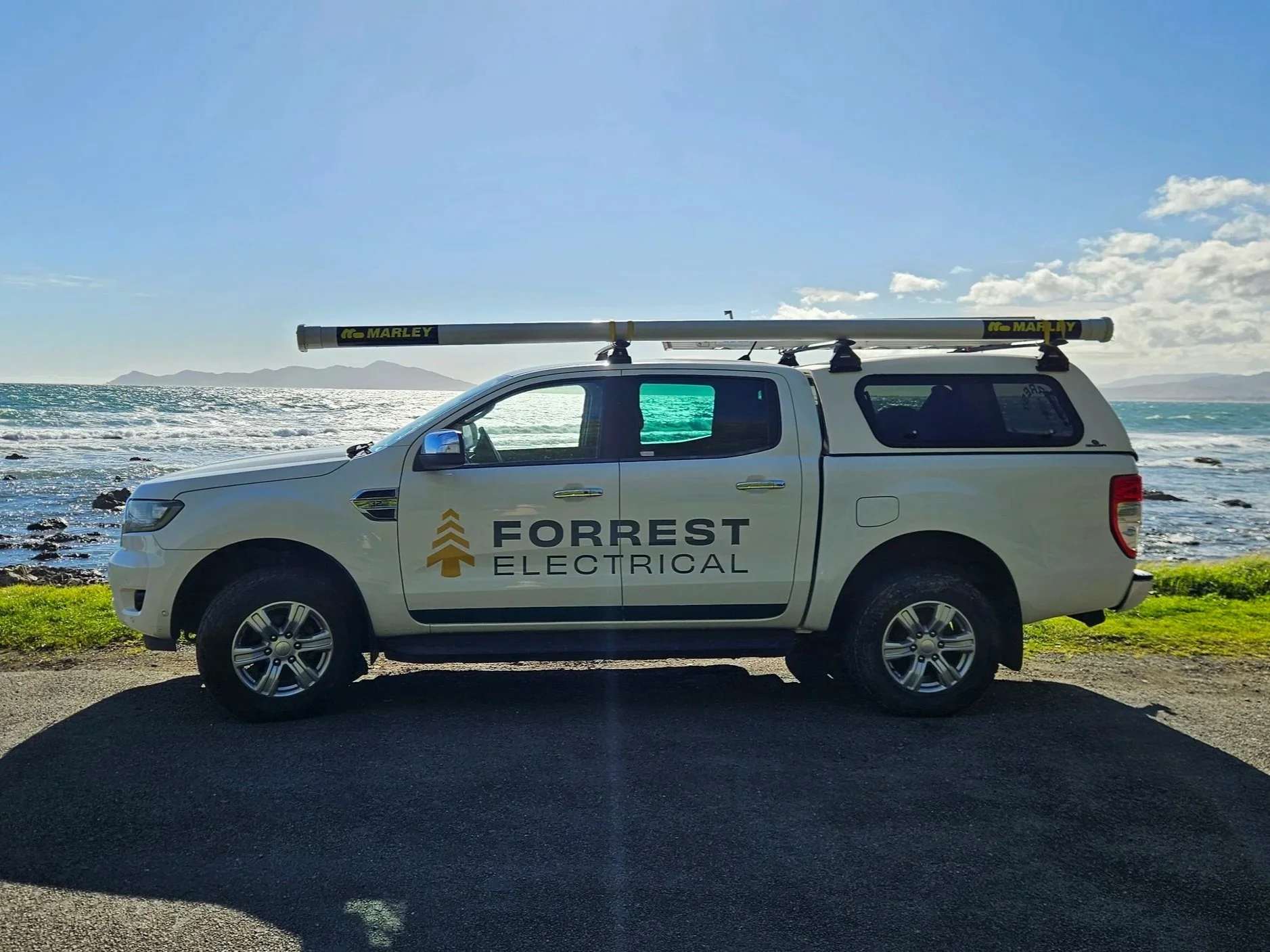 A white Ford pickup truck with 'Forrest Electrical' logo on the door, a yellow and black leveled pole on top, parked near the coast with ocean and mountains in the background, sunny sky, and some clouds.