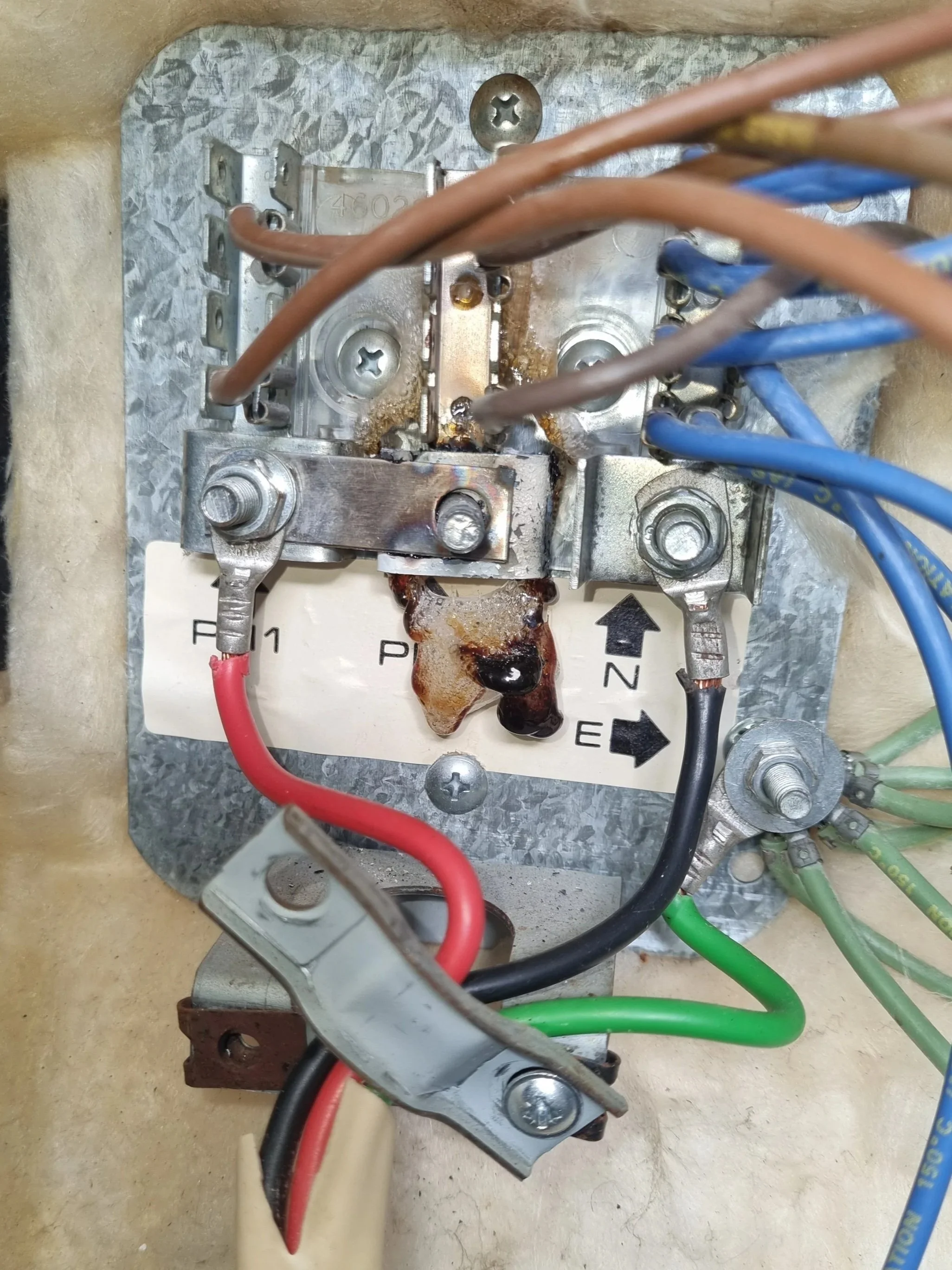 Close-up of an electrical circuit breaker with multiple wires connected to it, including red, black, green, and blue wires, mounted on a metal panel with screws and labels.