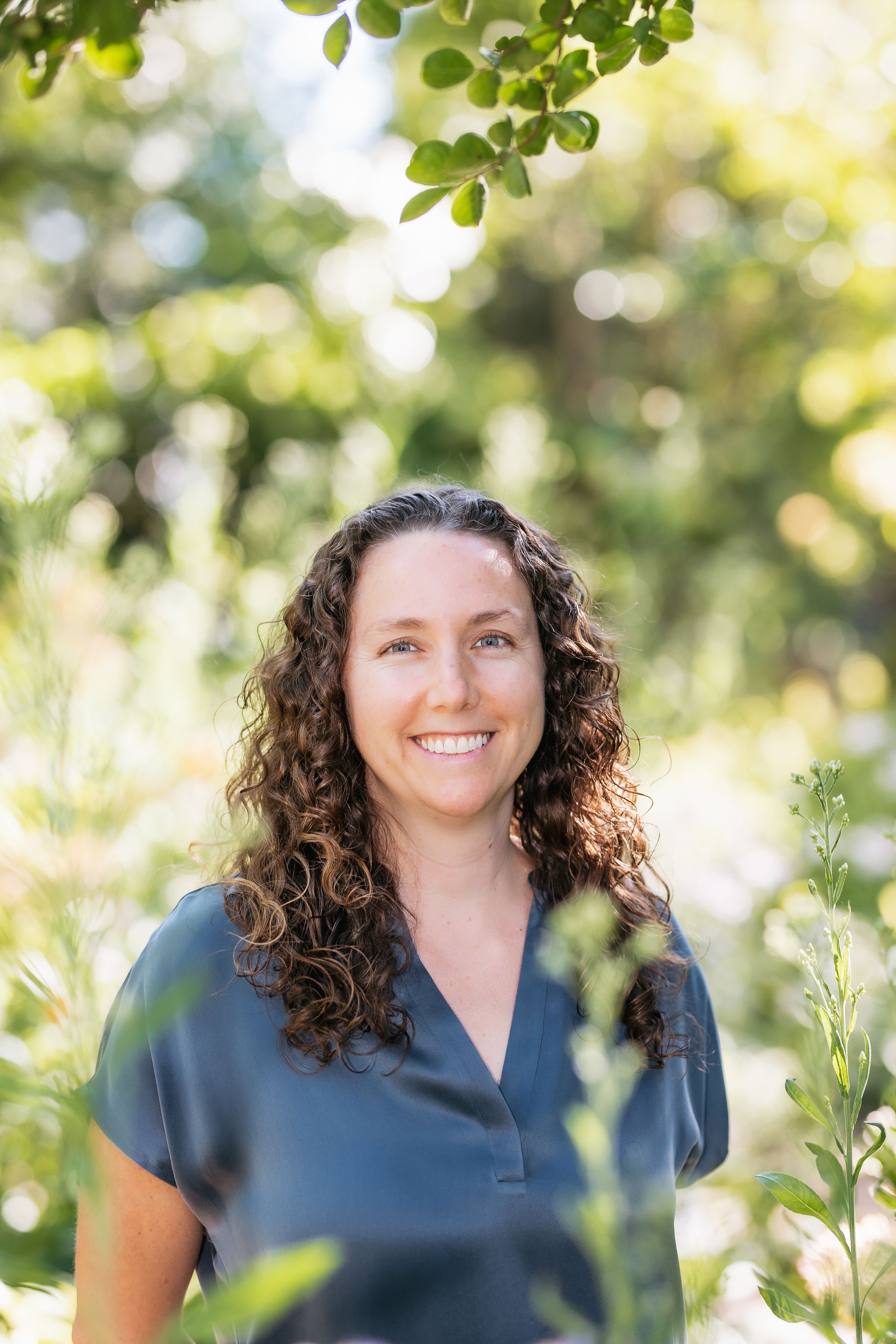 Woman with curly brown hair smiling at the camera sorrounded by green trees and plants