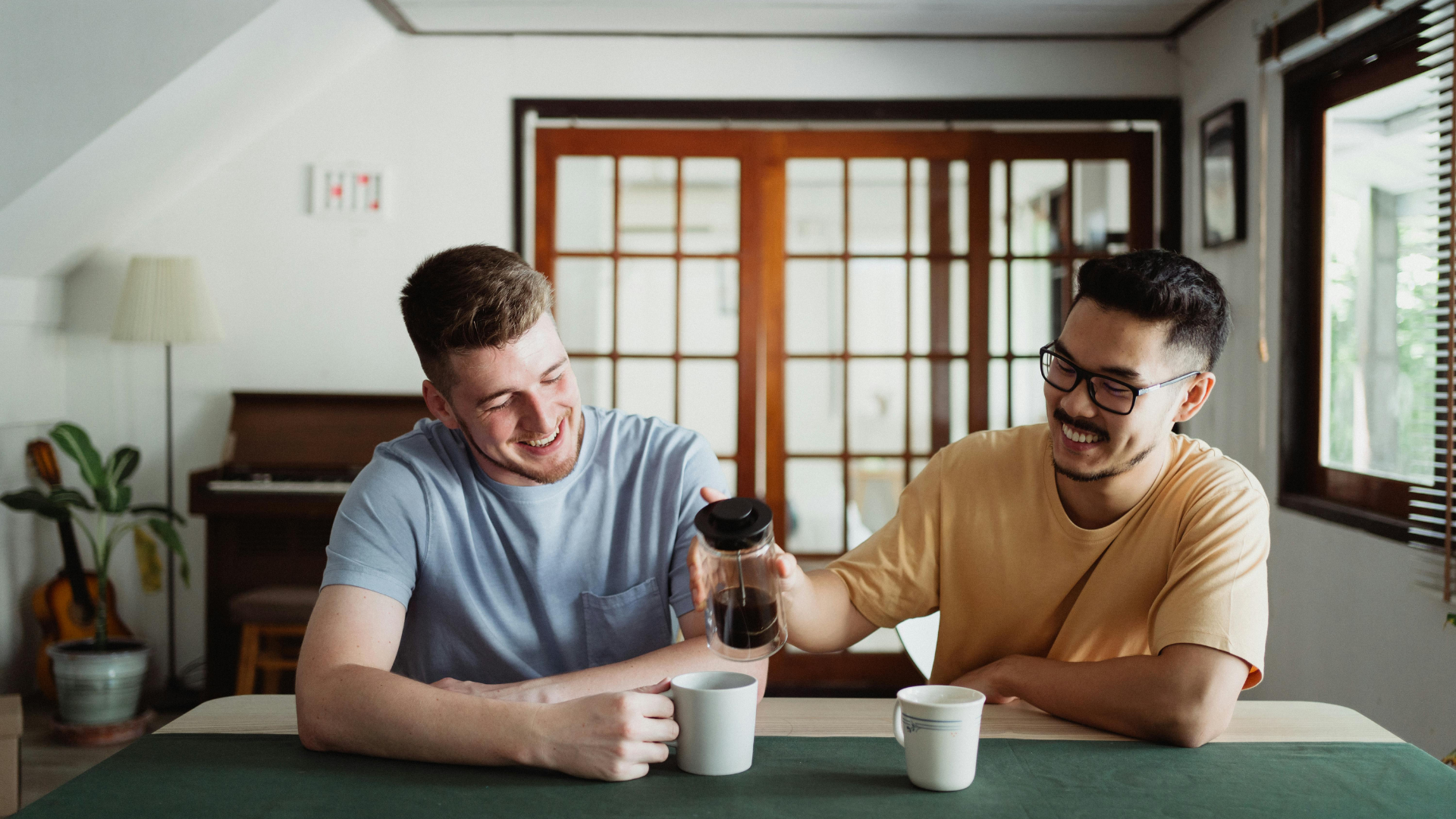 Two young men sitting at a table, one is pouring coffee into a mug, both smiling and enjoying each other's company in a cozy room with wooden accents and natural light.
