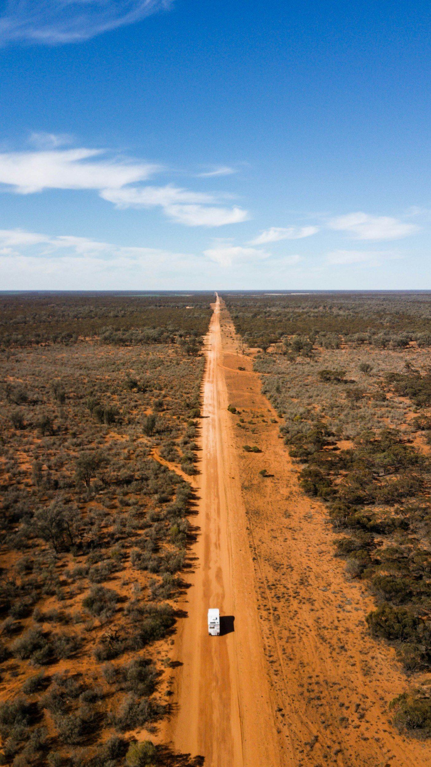 An aerial view of a long, straight dirt road stretching through a flat, arid landscape with sparse vegetation and a clear blue sky overhead.