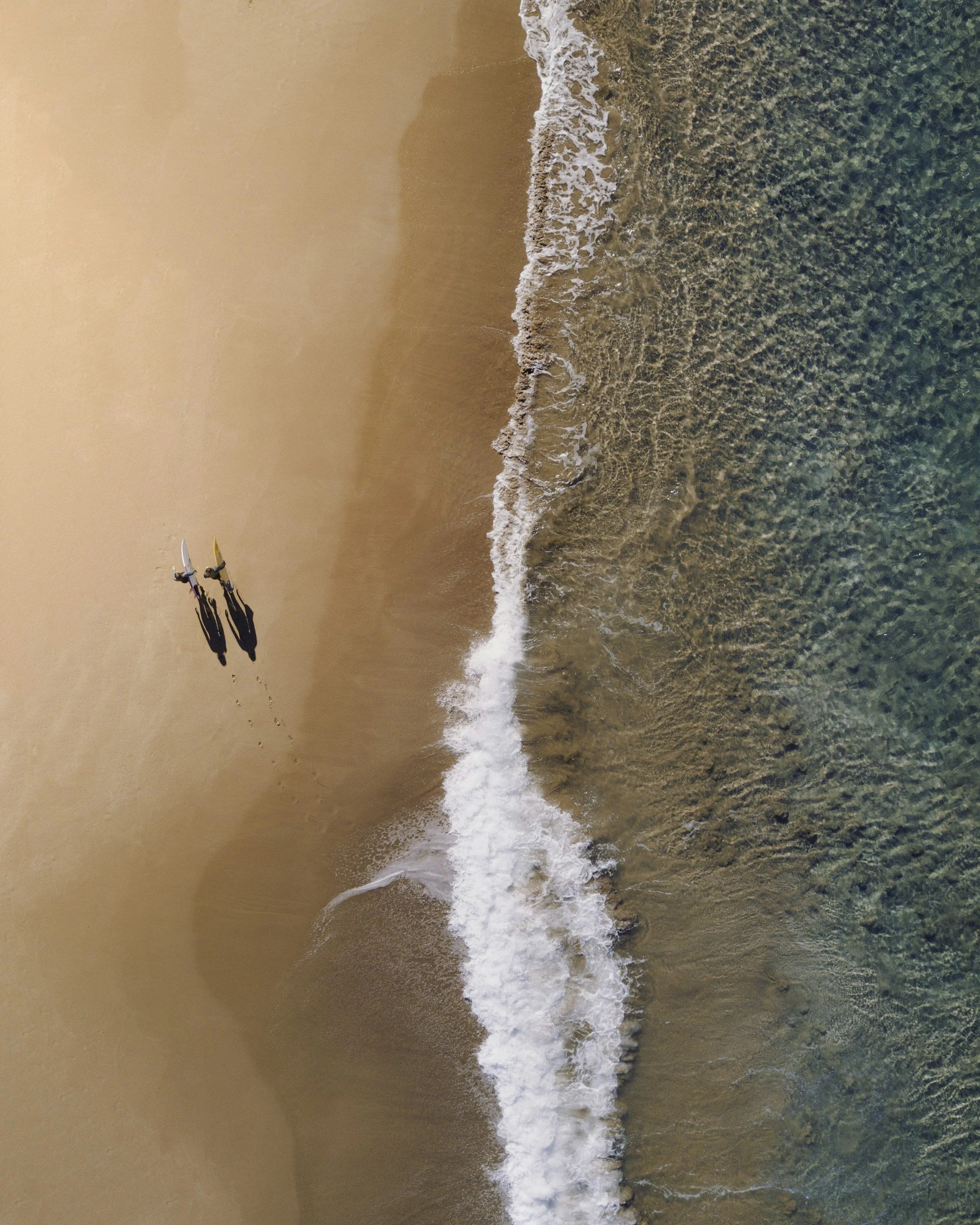 Two surfers walking on a sandy beach near the water with waves crashing in.
