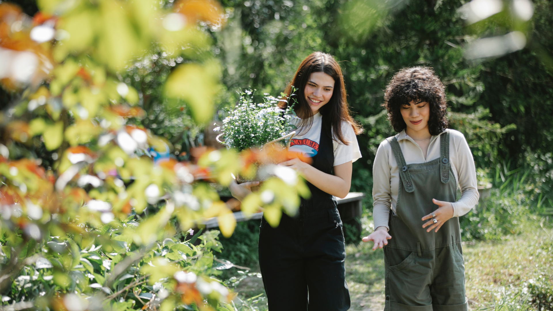 Two women in a garden, one holding a potted plant and both smiling, surrounded by green plants.