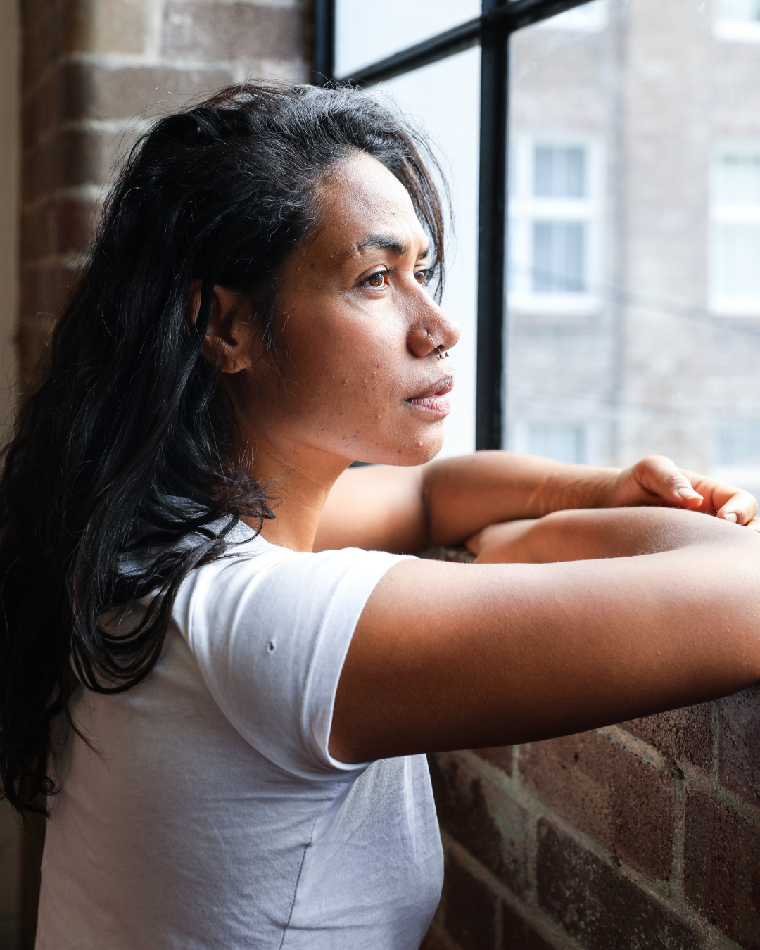 A woman with dark hair and a nose ring looking out of a window, resting her arms on the windowsill, in a room with brick walls.