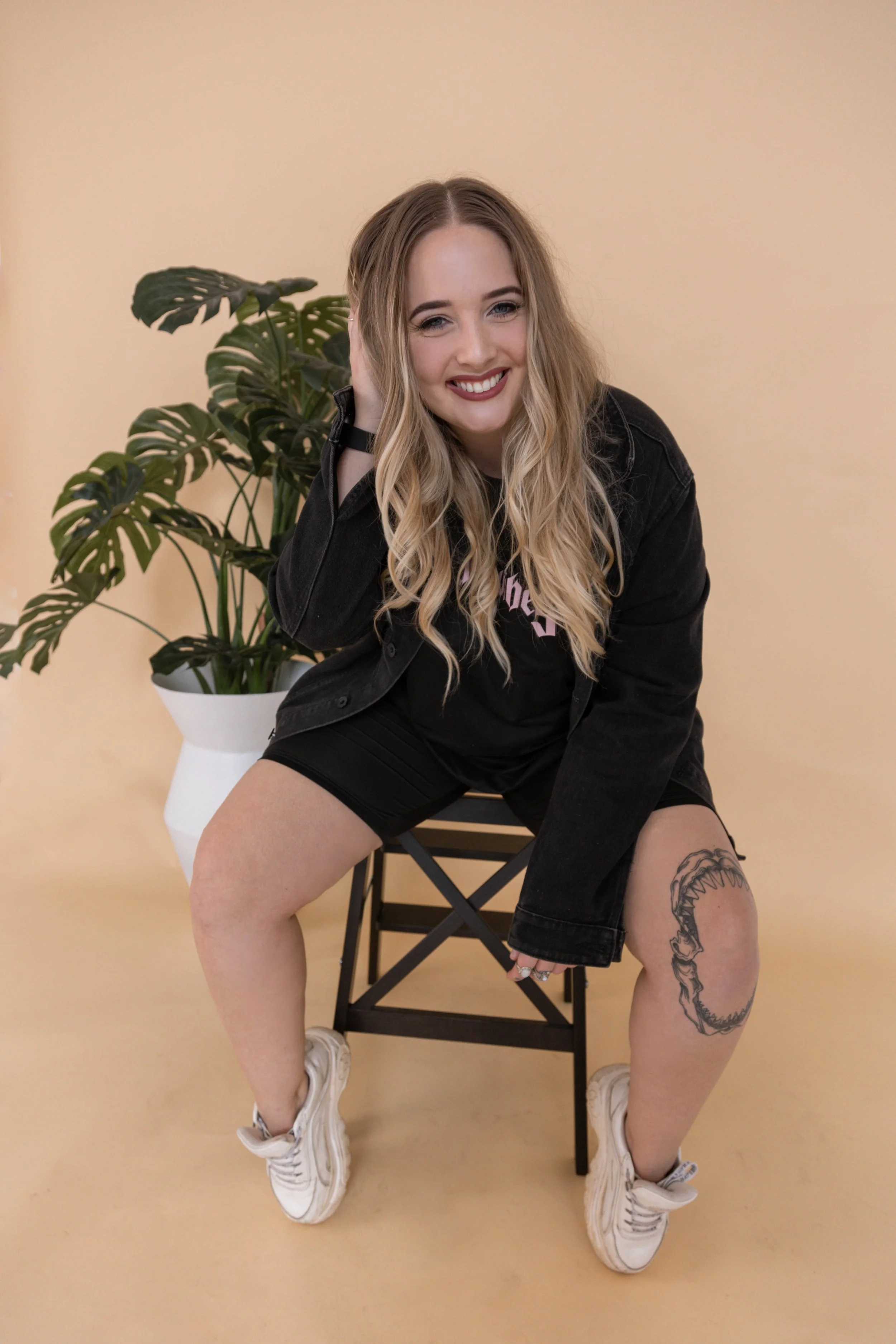 A woman with long wavy blonde hair sitting on a black chair, smiling, wearing a black jacket, black shorts, and white sneakers, with a tattoo of a shark on her left thigh, in front of a yellow wall and a large potted plant.