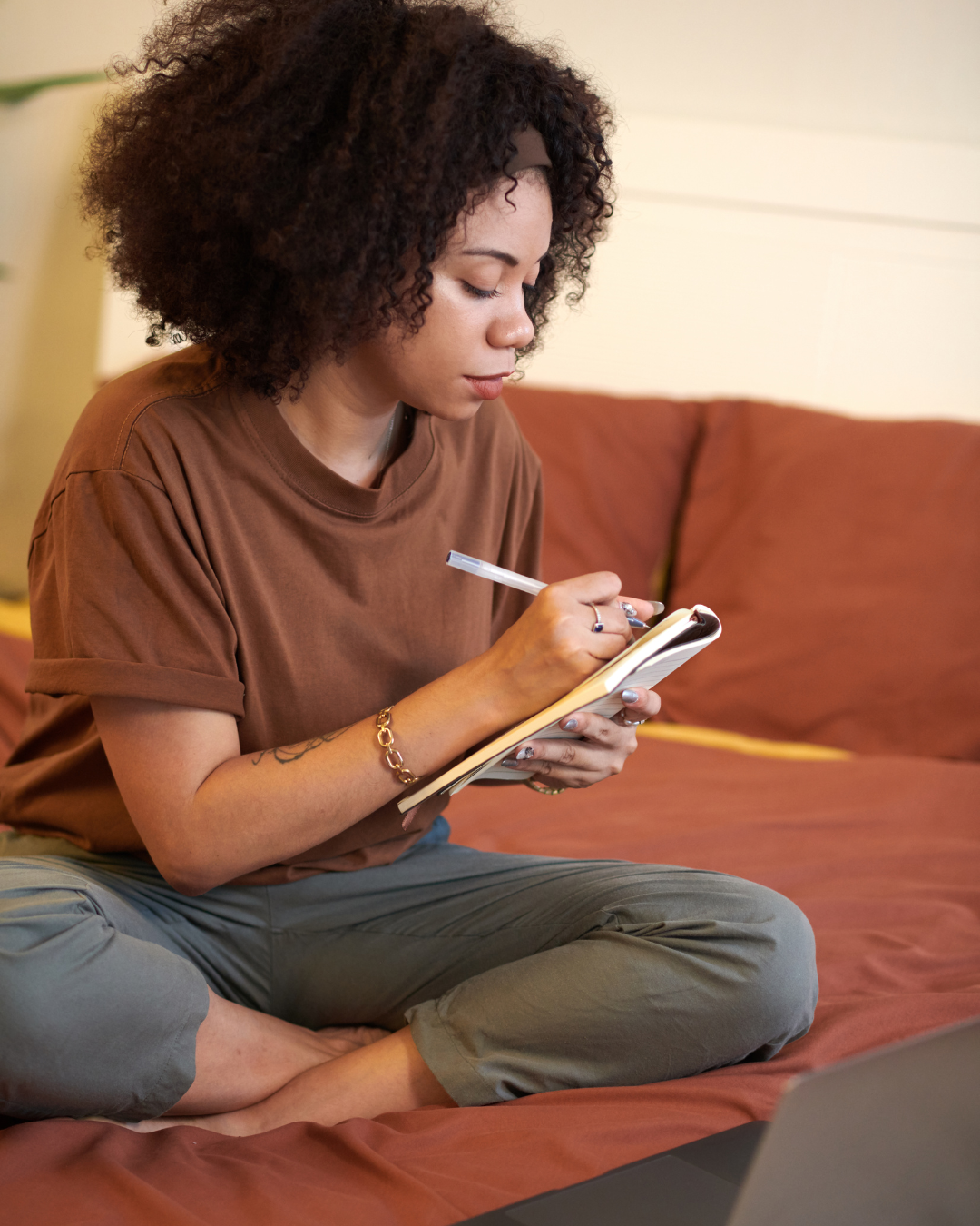 A woman with curly hair, sitting cross-legged on a couch, writes in a small notebook with a pen, with a laptop in front of her.