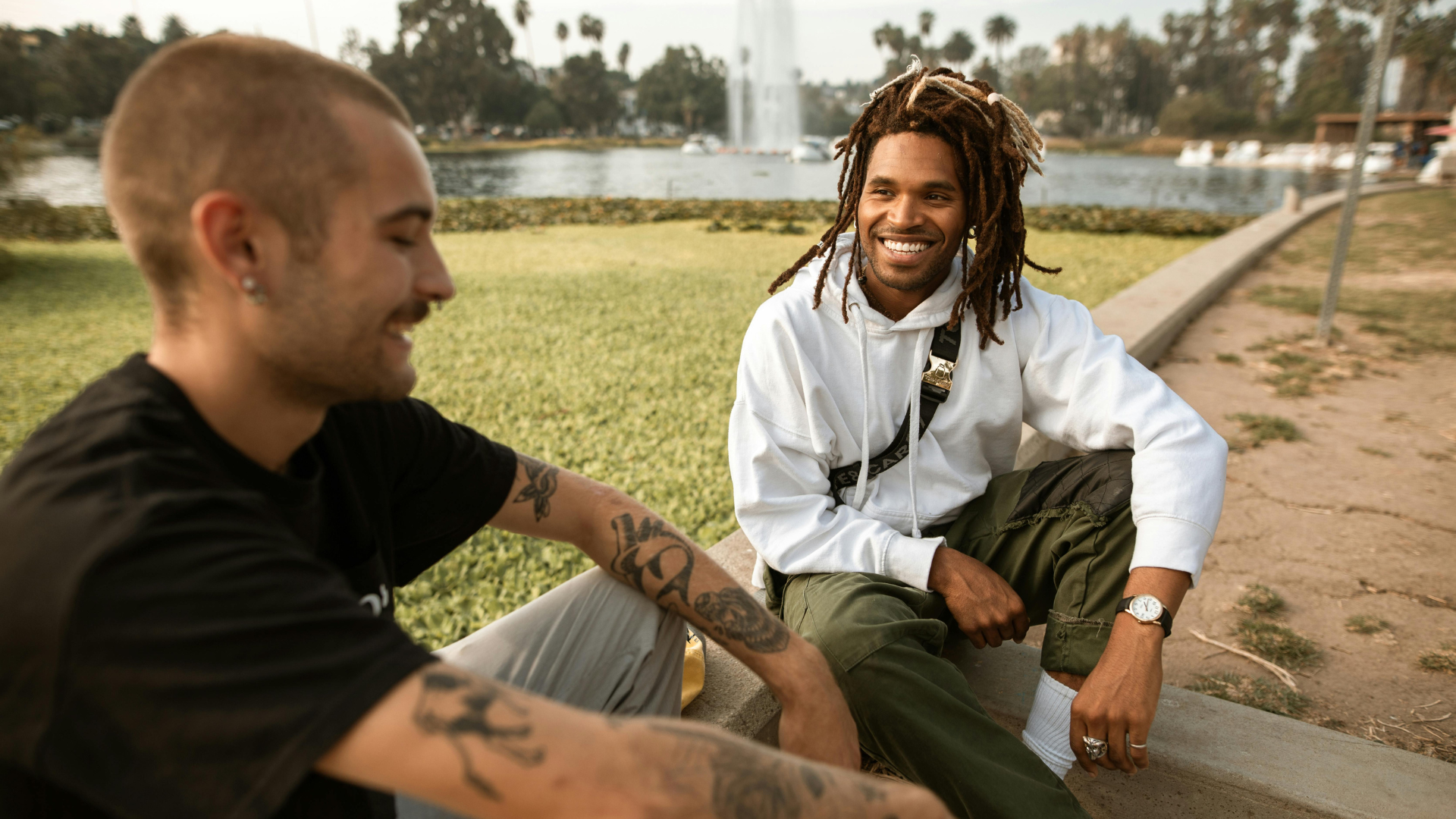 Two young men sitting outdoors by a lake, smiling and talking, with trees and water fountain in the background.