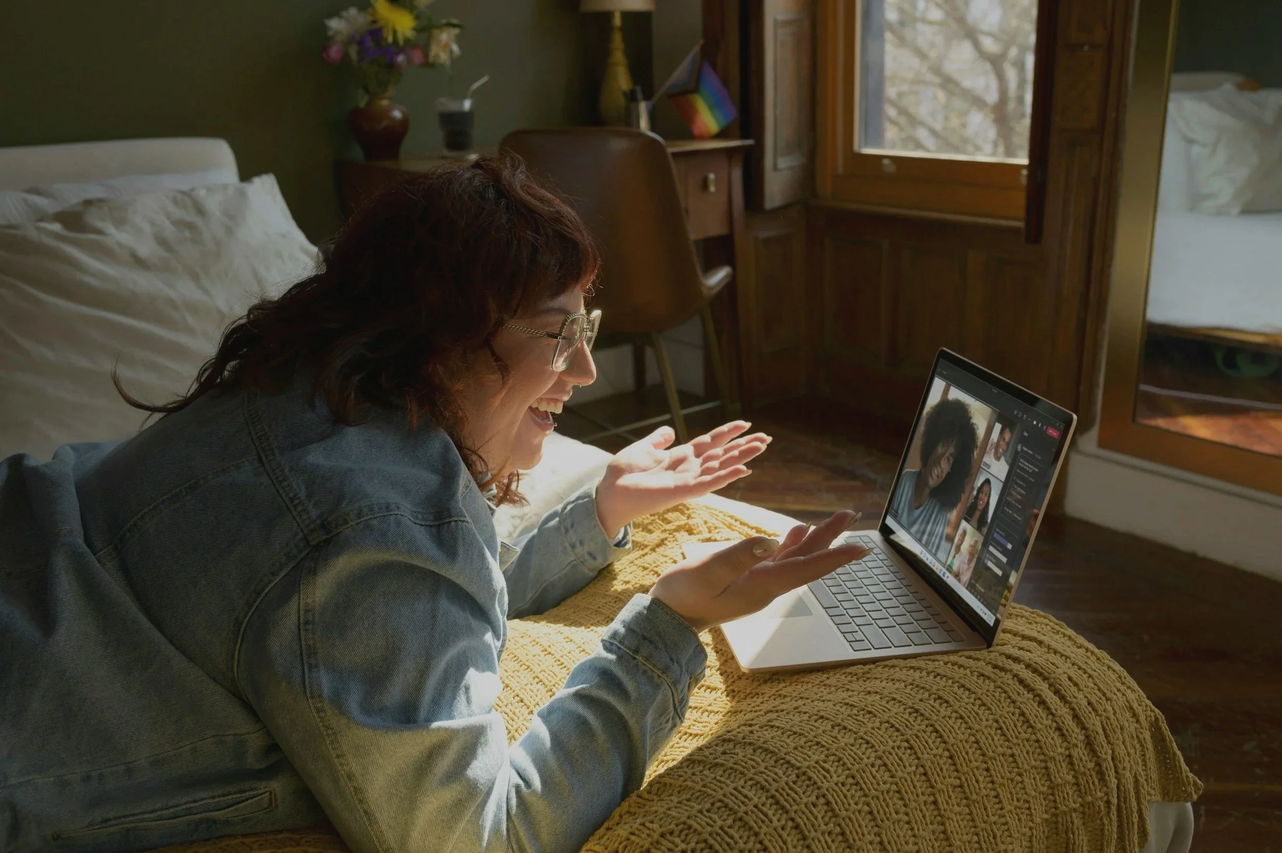 A woman with curly hair, glasses, and a denim jacket lying on her stomach on a bed, smiling and waving at her laptop during a video call in a cozy room with wooden furniture and a window.