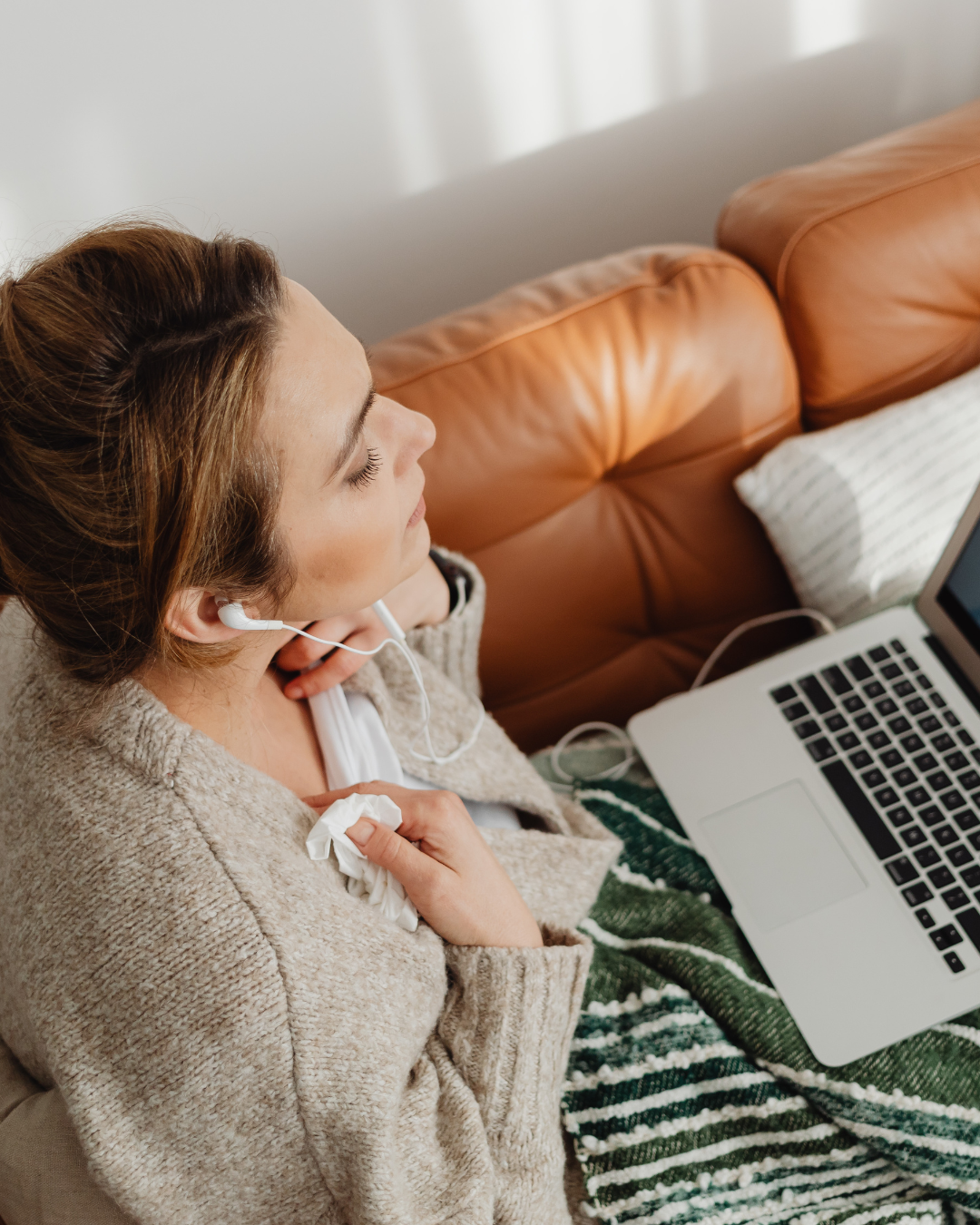 Woman with earbuds sitting on a couch, holding tissue, in front of a laptop.