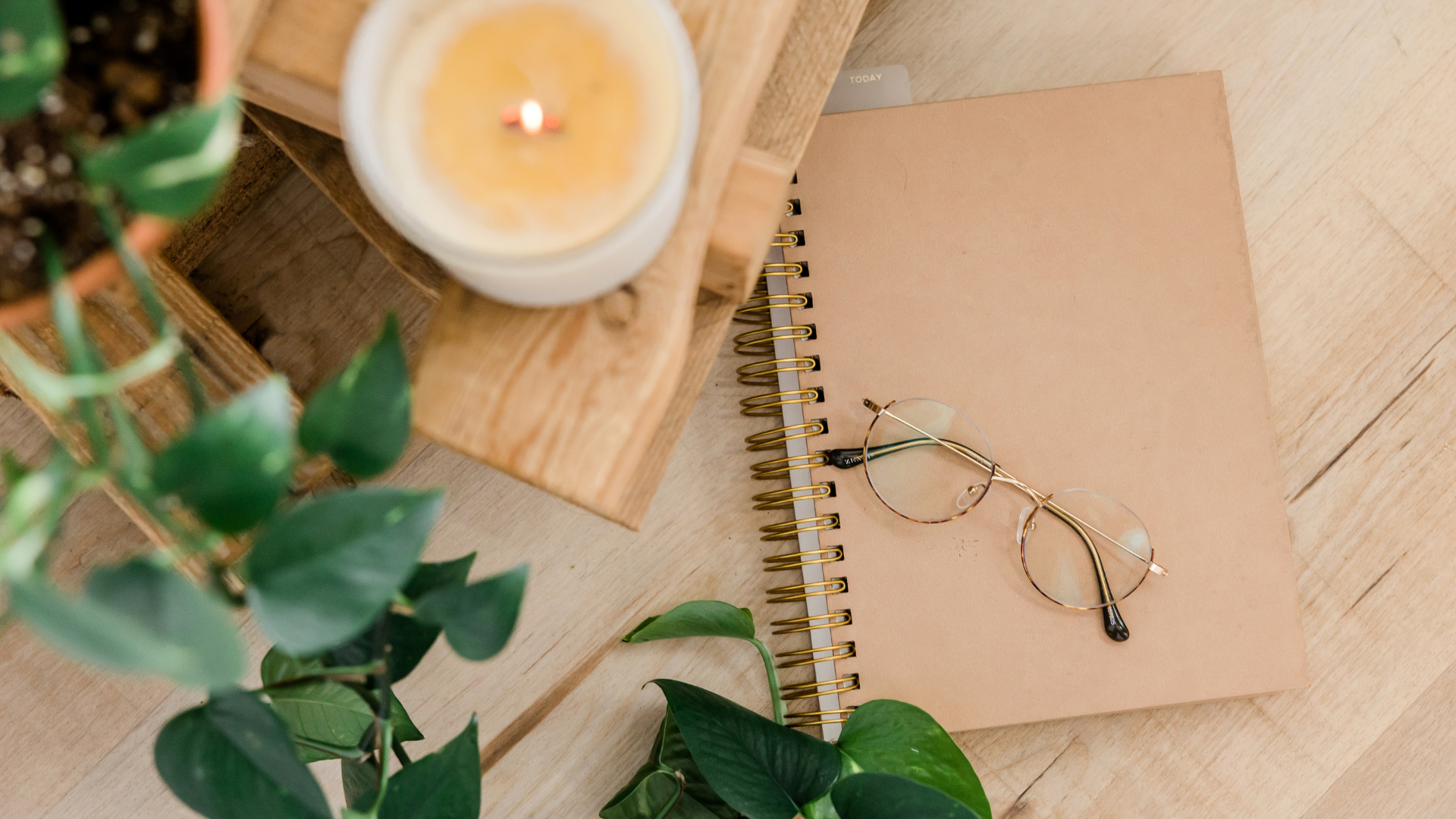 A close-up of a desk with a spiral notebook, a pair of glasses, a candle, and some green plants on a wooden surface.
