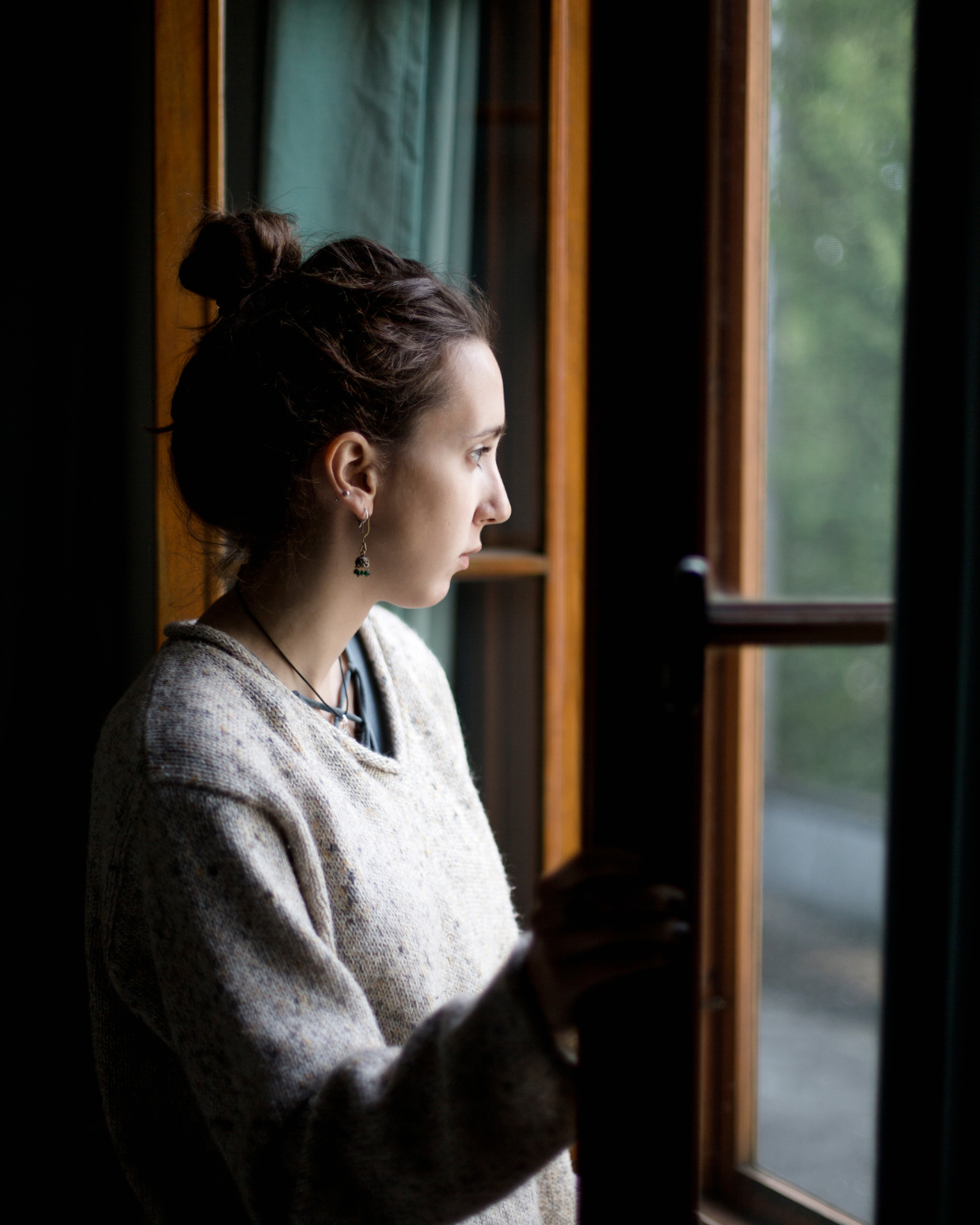A young woman with dark hair in a bun, wearing earrings, looking out a window.