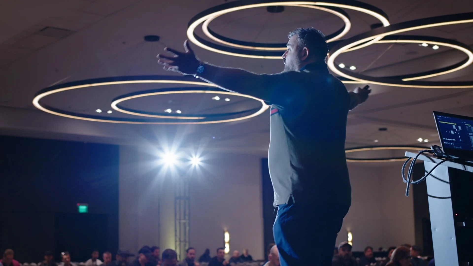 A speaker on stage at an Austin conference or seminar, gesturing with arms extended, with an audience in the background under modern circular ceiling lights. Another still image by the Austin Videographer.