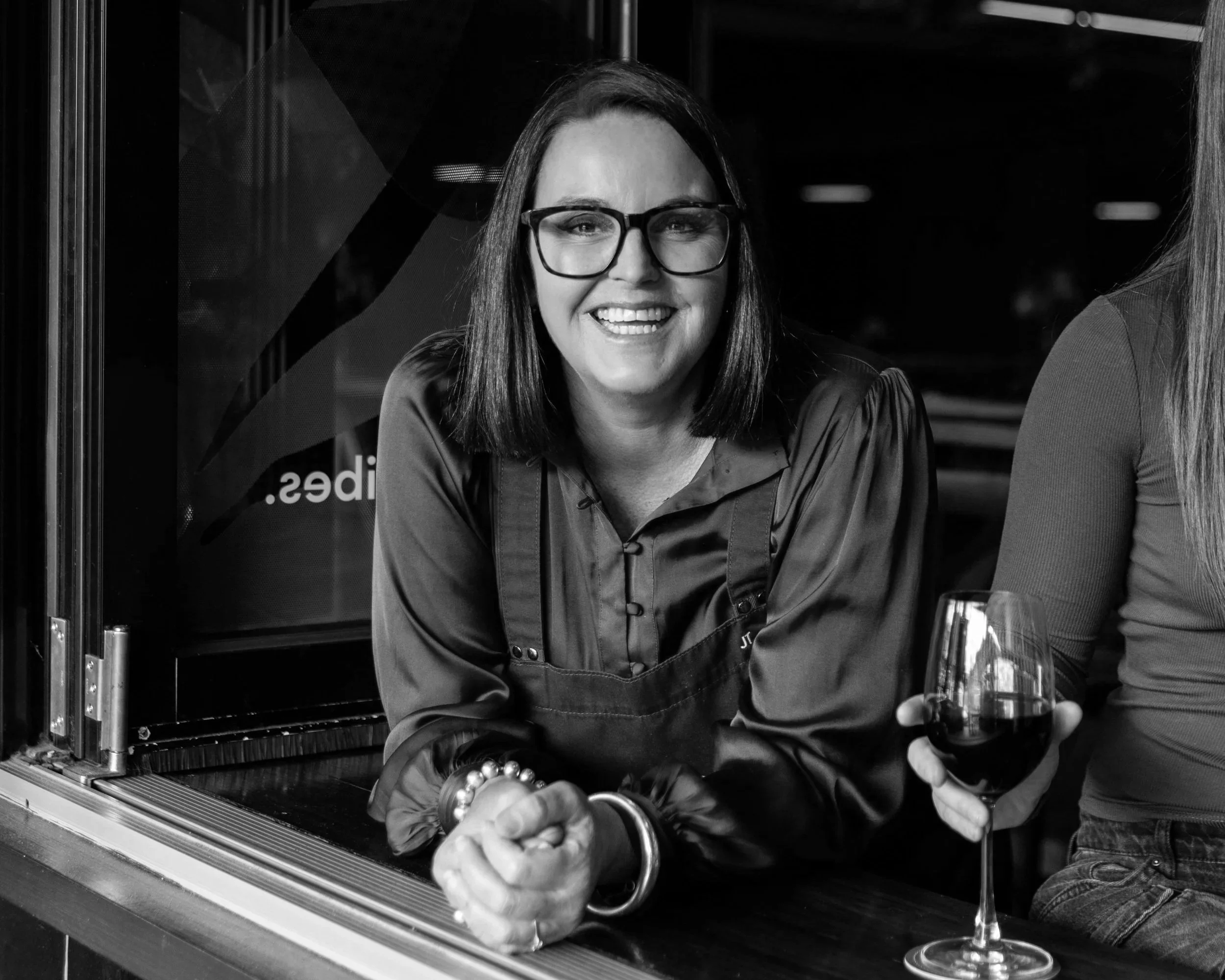 A woman with glasses smiling at a bar, holding a glass of red wine.