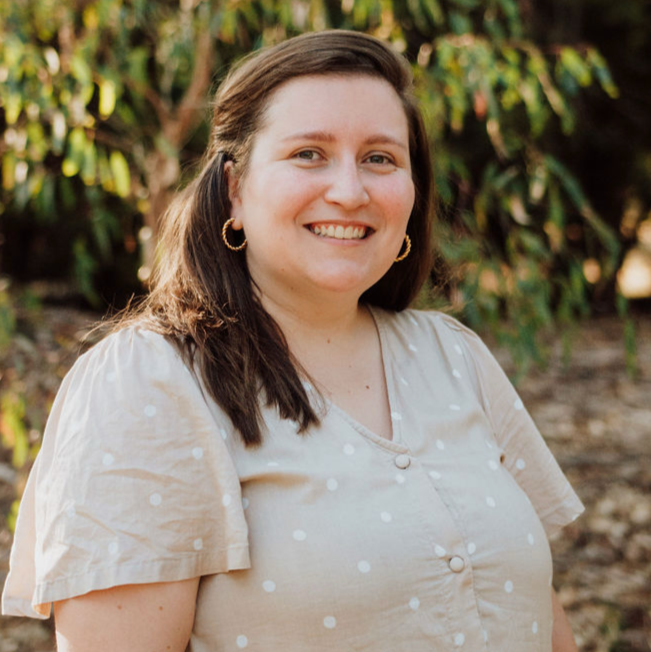 Smiling woman with shoulder-length dark hair, wearing hoop earrings and a beige dress with white polka dots, outdoors with green foliage background.