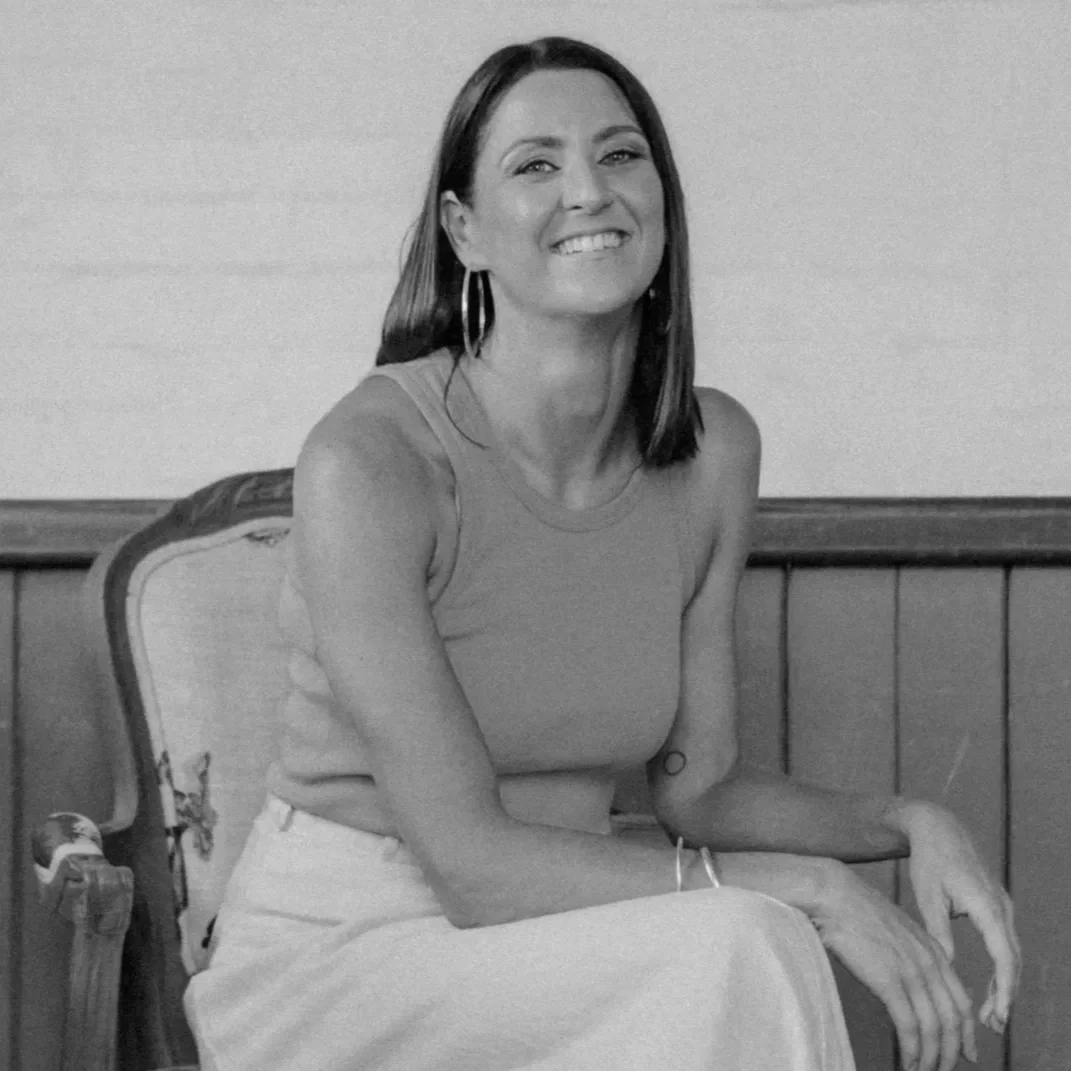 A woman with shoulder-length hair, wearing large hoop earrings, a sleeveless top, and light-colored pants, sitting on a wooden chair with a smile.