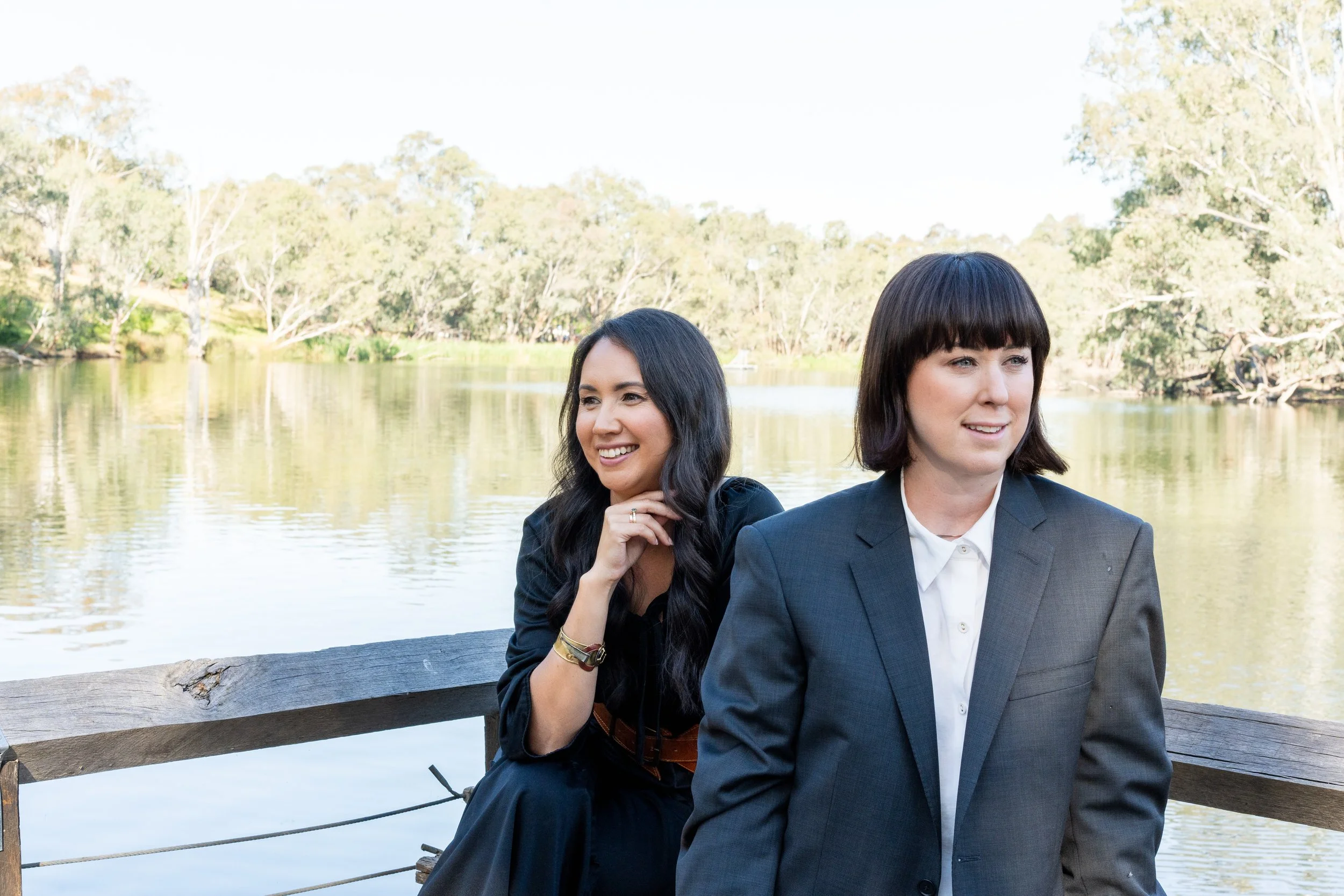 Two women sitting near a lake with trees in the background, one in a black dress smiling, the other in a gray suit looking away.
