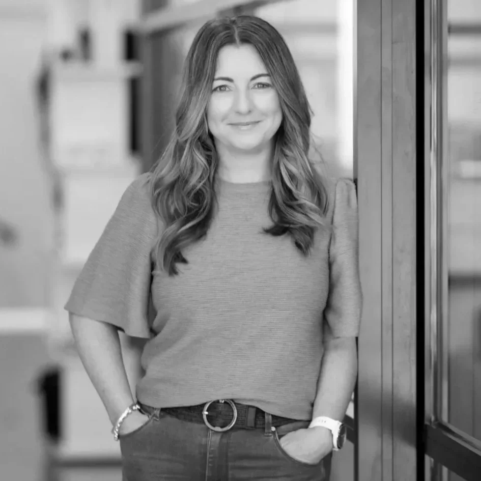 A woman with wavy hair wearing a short-sleeved shirt and jeans, standing indoors with her hands in her pockets, smiling at the camera.