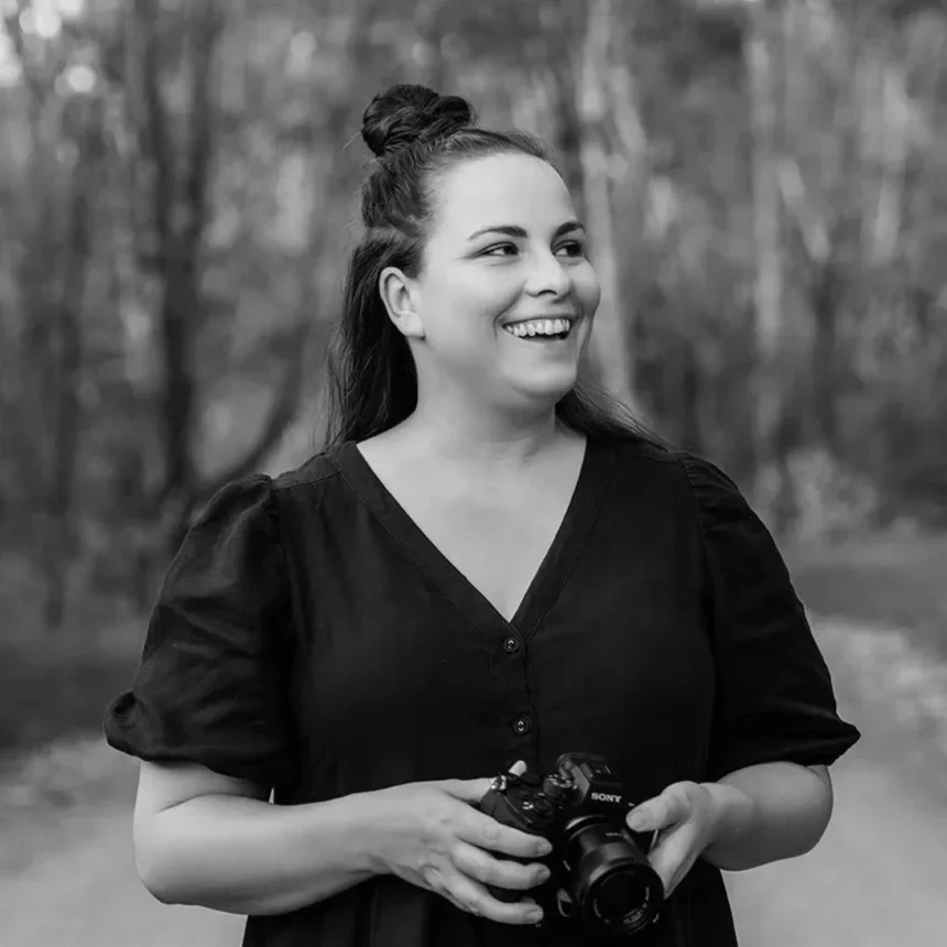 A woman smiling outdoors holding a camera, with trees in the background.