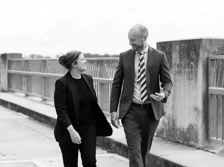 Two business professionals, a woman and a man, are walking and talking outdoors on a bridge, smiling and engaging in conversation.