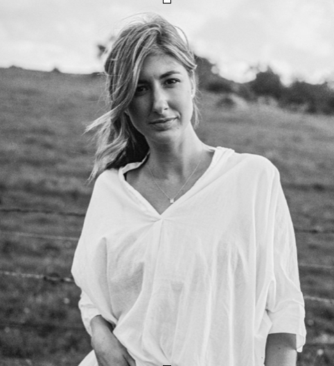 Black and white photo of a woman standing outdoors with a grassy field in the background. She has shoulder-length hair, slightly tousled, and is wearing a loose, light-colored top.