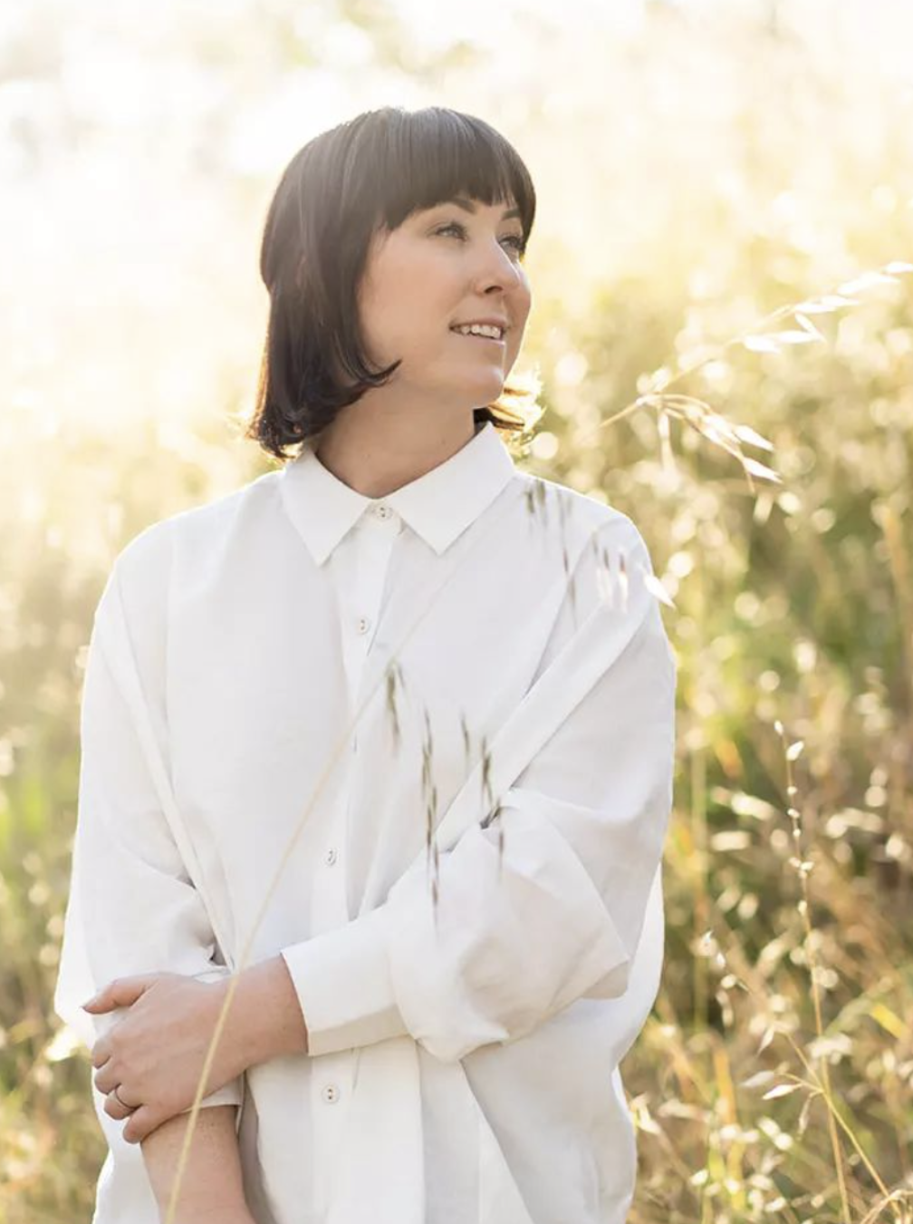 A woman with short dark hair wearing a white button-up shirt standing outdoors in a field with tall grass and plants, illuminated by sunlight.