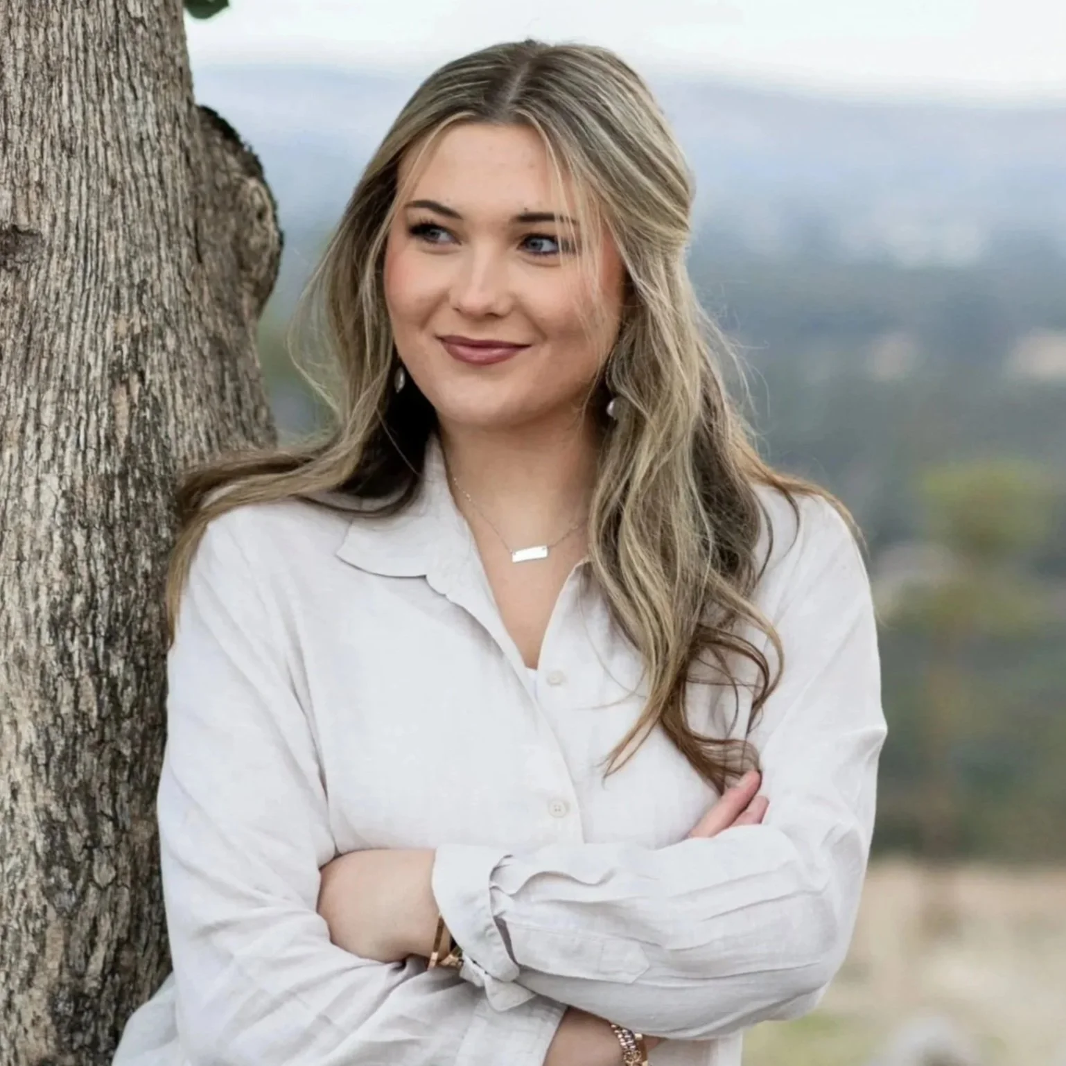 A young woman with wavy blonde hair, wearing a white button-up shirt, standing outdoors next to a tree with mountains in the background.