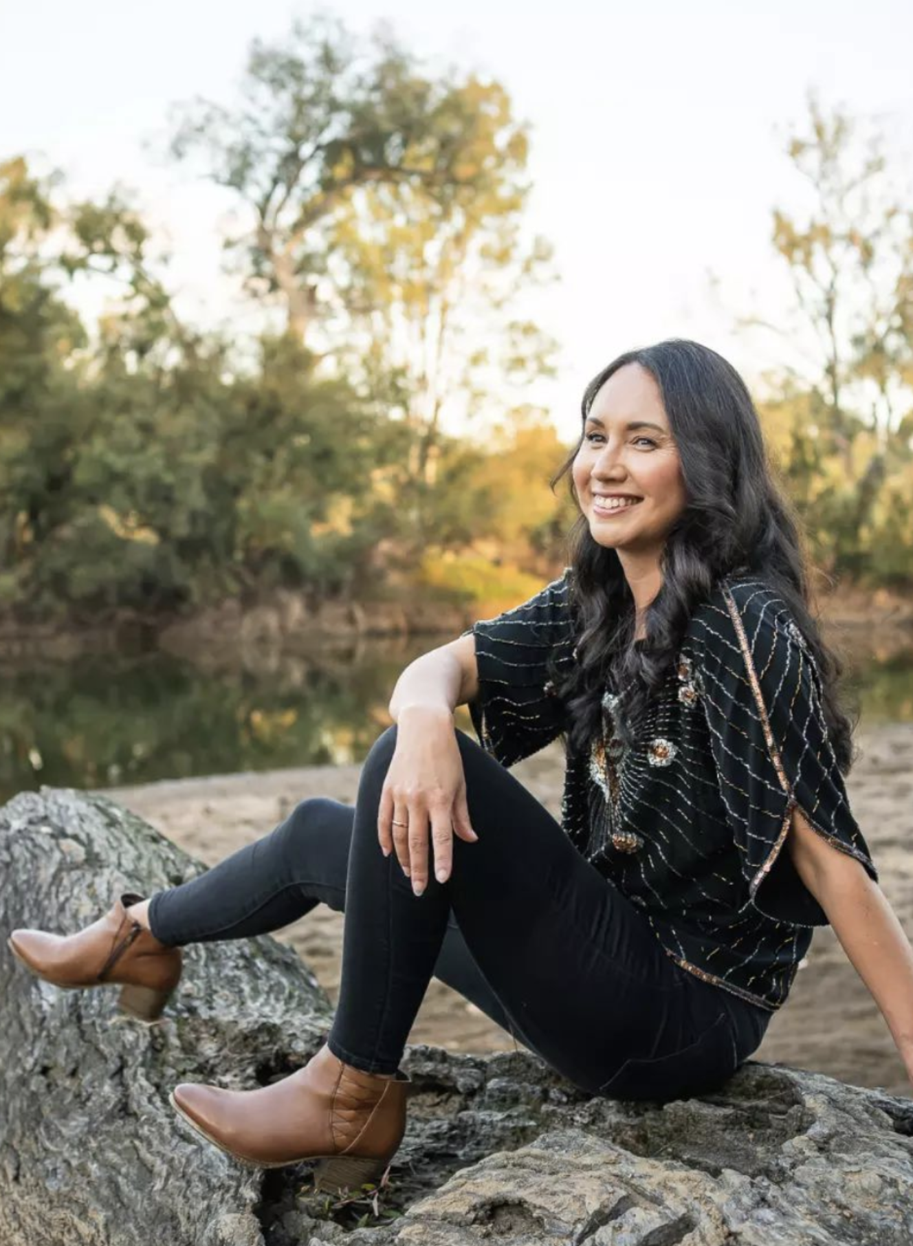 A smiling woman with long dark hair sitting on a large tree log by a river, wearing a black patterned top, black jeans, and brown ankle boots, during sunset with trees in the background.