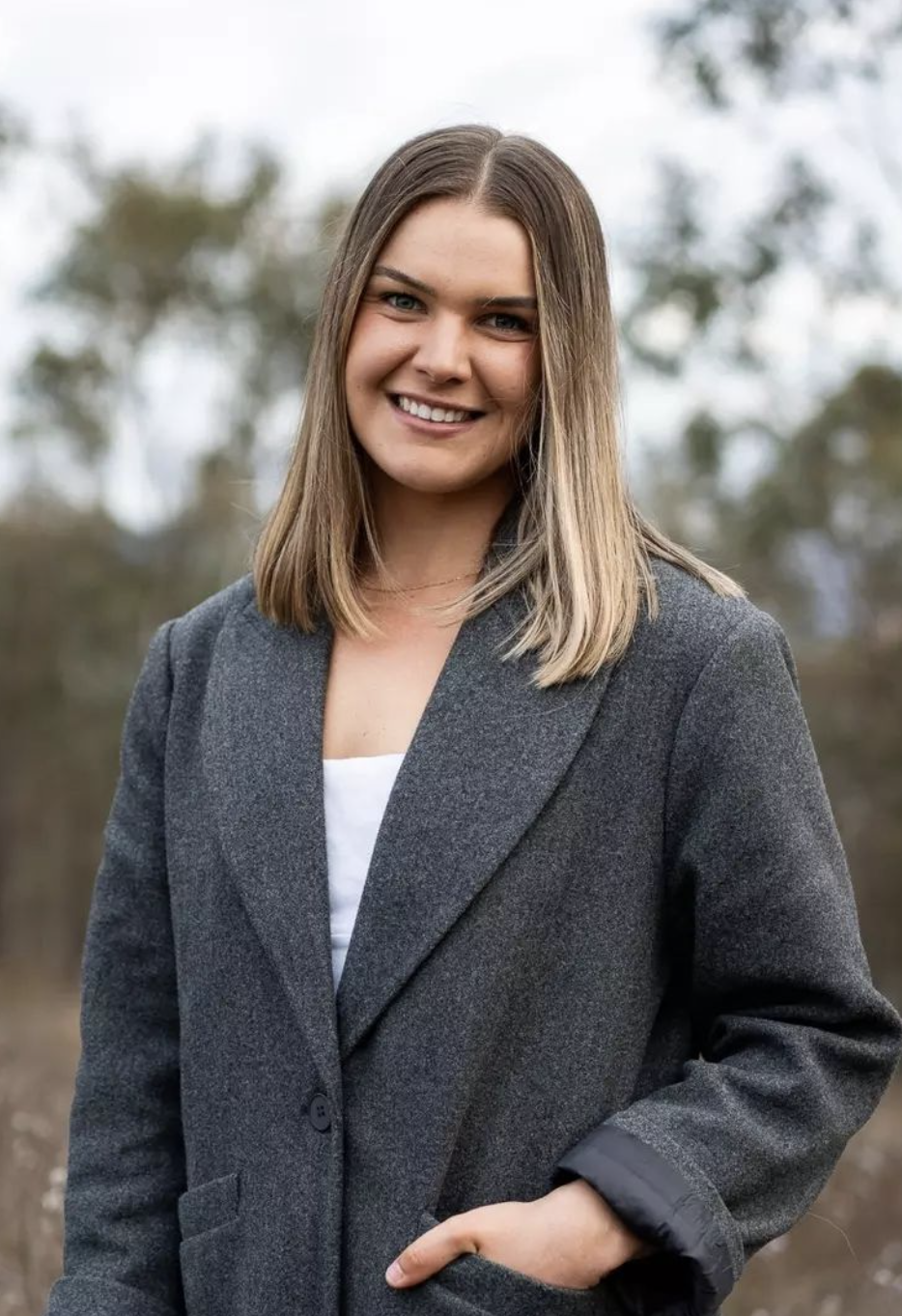 A smiling young woman with shoulder-length brown hair, wearing a gray blazer over a white top, standing outdoors with blurred trees in the background.
