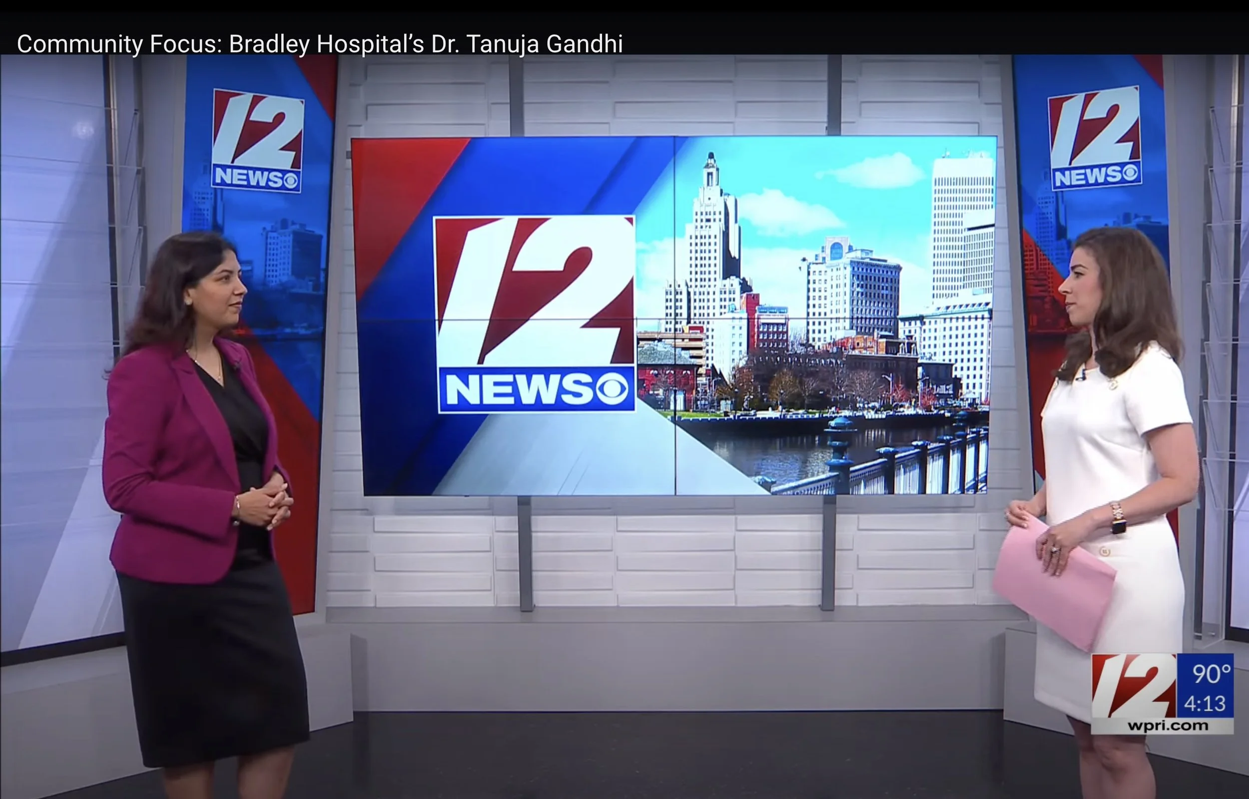 Two women are standing in a news studio, facing each other, with a large digital screen behind them displaying the '12 News' logo and a cityscape of Providence, Rhode Island. One woman is wearing a magenta blazer and black dress, and the other is in a white dress holding a pink folder.
