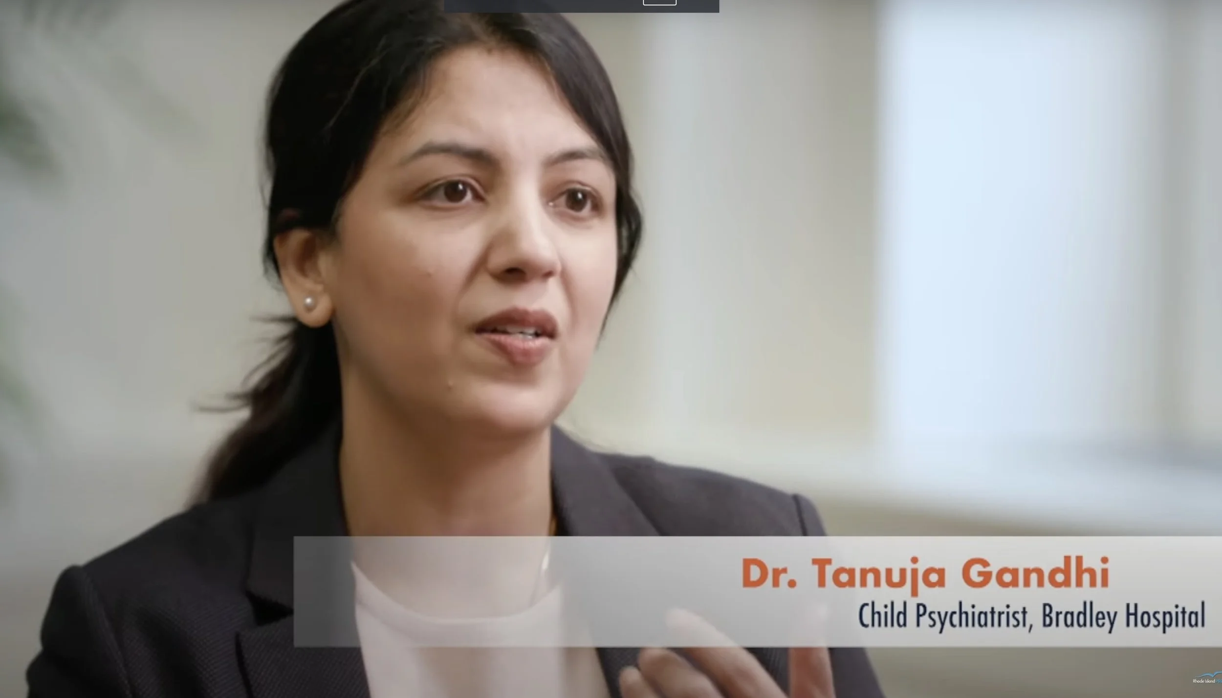 A woman with dark hair, wearing a black blazer and pearl earrings, is speaking during an interview. There is a text overlay showing her name, Dr. Tanuja Gandhi, and her title, Child Psychiatrist at Bradley Hospital.