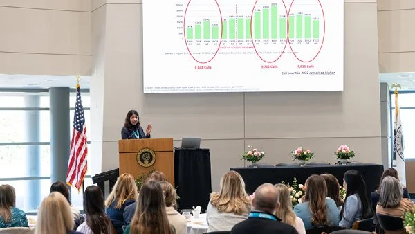 A woman giving a presentation at a conference, standing behind a podium with an American flag beside her. The backdrop features a large screen displaying a bar graph and data points. Audience members sit facing her.