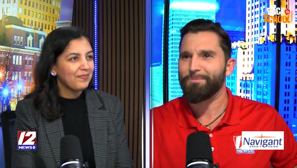 Two people, a woman and a man, are speaking into microphones during a news interview, with a cityscape background visible through large windows.