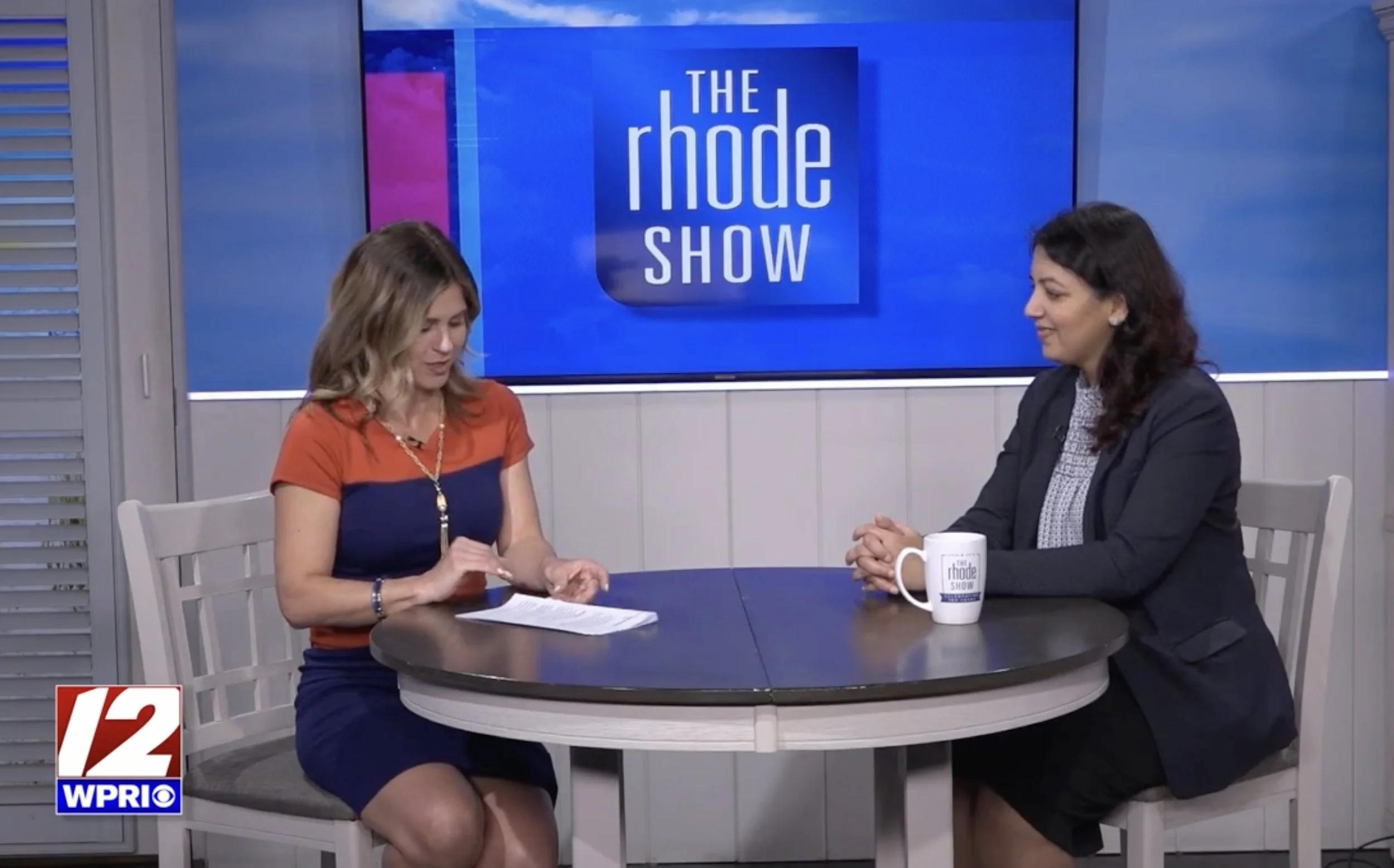 Two women having a conversation on the set of a TV show called 'The Rhode Show.' They are sitting at a round table, with one woman reading notes and the other listening, with a "WPRI" logo in the bottom left corner.