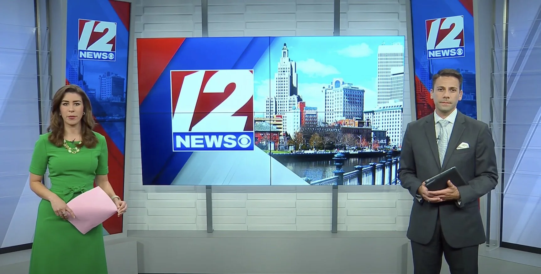 Two news anchors, a woman in a green dress holding pink papers and a man in a gray suit holding a tablet, stand in a TV studio with a large screen showing the 12 News logo and a cityscape in the background.