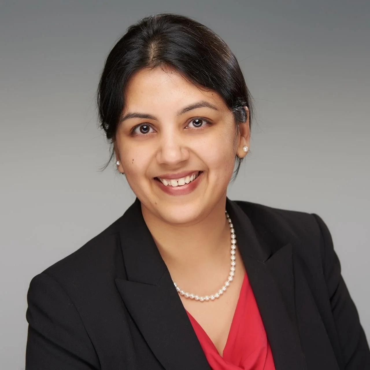 Professional woman wearing a black blazer, red blouse, pearl necklace, and pearl earrings, with short dark hair and a smile, against a gray background.