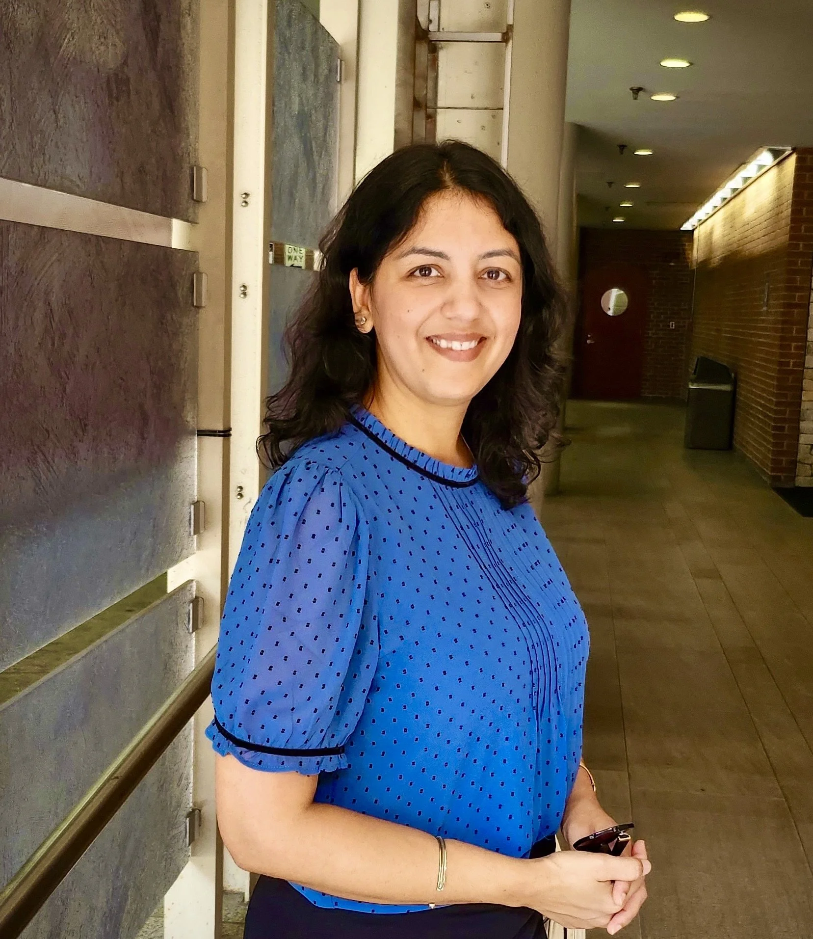 A woman with black hair wearing a blue polka dot blouse, smiling and standing in a hallway.