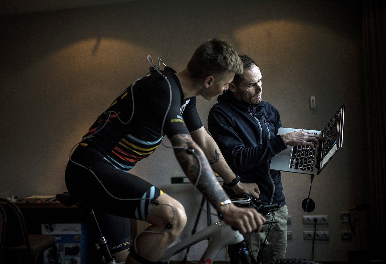 A cyclist on a stationary bike being monitored and analyzed by a researcher with a laptop, during a lab test.
