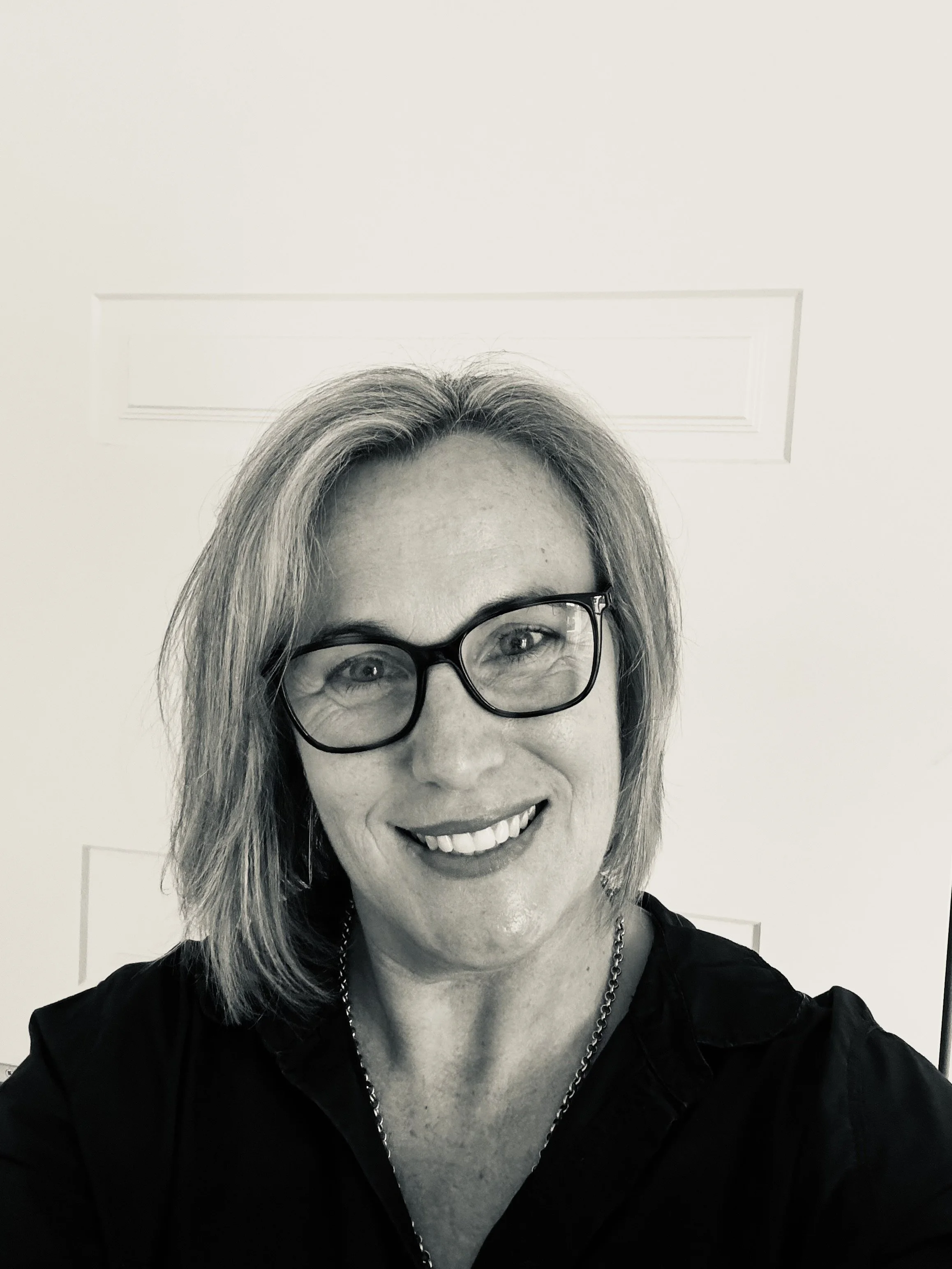 A woman with shoulder-length hair, glasses, and a necklace smiling for a selfie in front of a plain door.