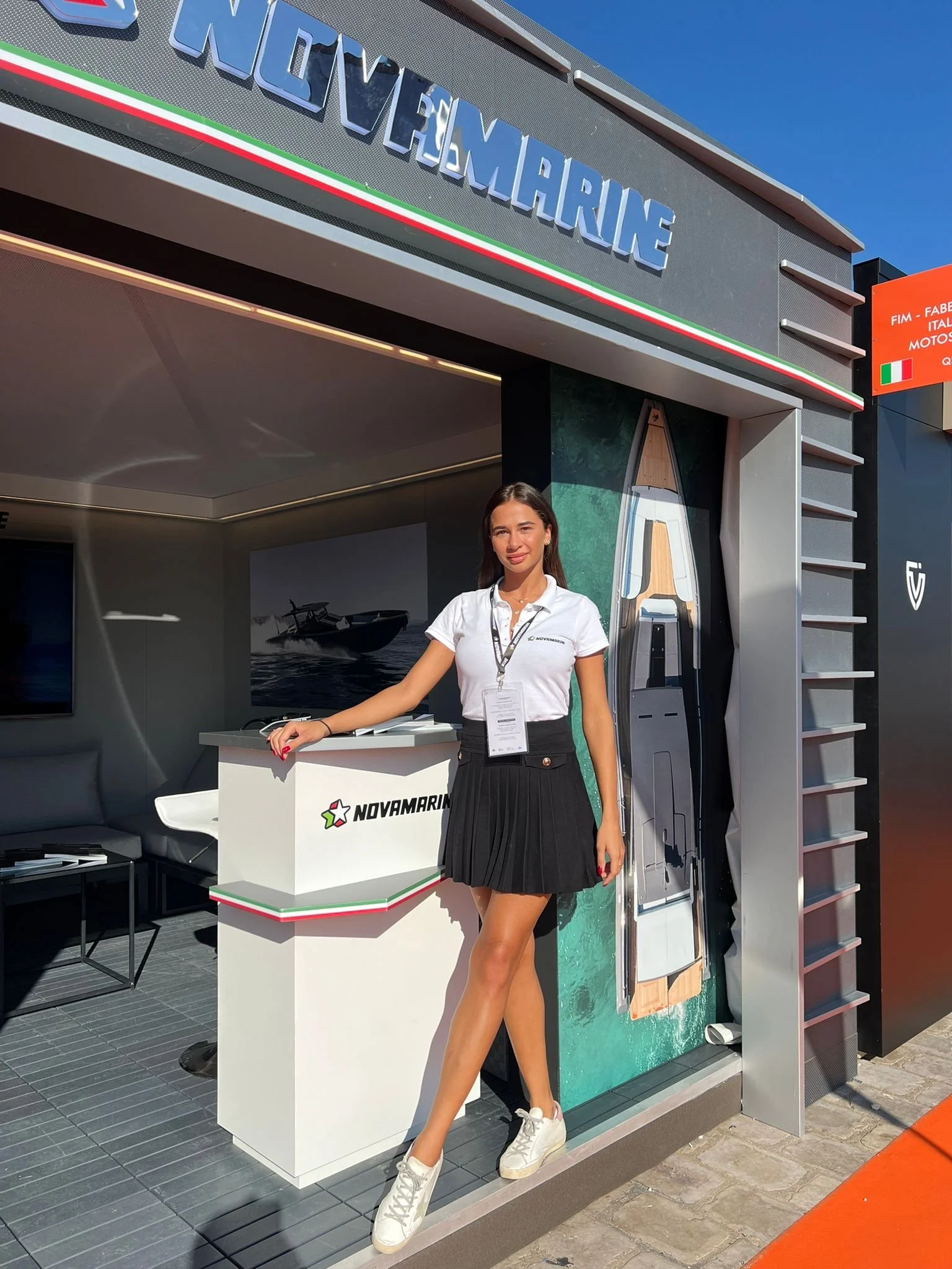 Una donna in abbigliamento casual con una maglietta bianca e una gonna nera davanti a uno stand Novamarine, una compagnia di imbarcazioni, con un display di una barca tra le mani.
