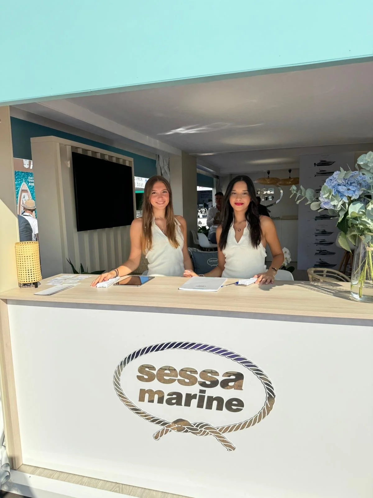 Due donne dietro il bancone di un evento con il logo 'Sessa Marine', decorazioni floreali e persone sullo sfondo.