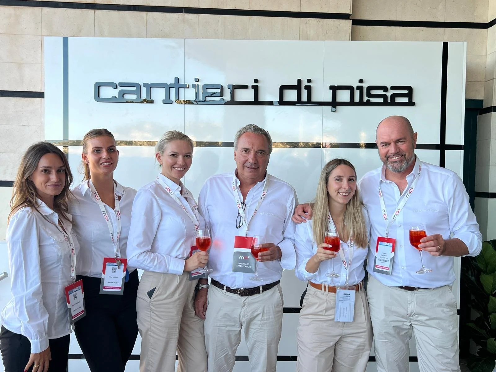 Gruppo di sei persone sorridenti in abiti bianchi, che tengono dei bicchieri con bevande rosse, davanti a un cartello che dice 'cantieri di pisa'
